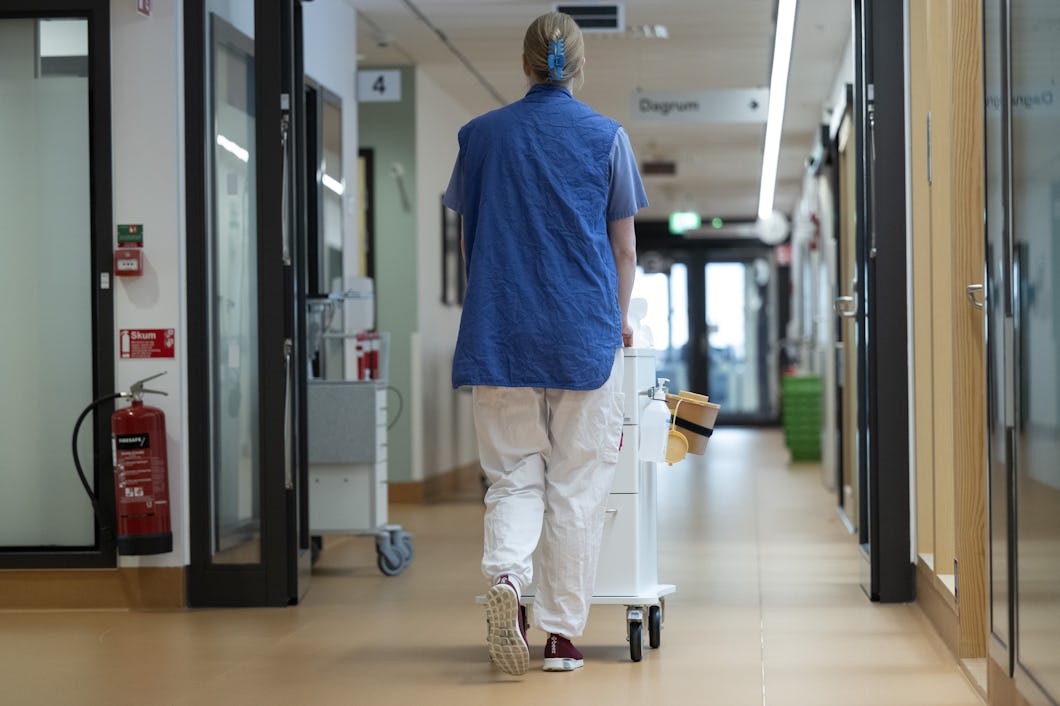 En vårdpersonal i arbetskläder och väst skjuter en medicinvagn genom en sjukhuskorridor, sedd bakifrån.