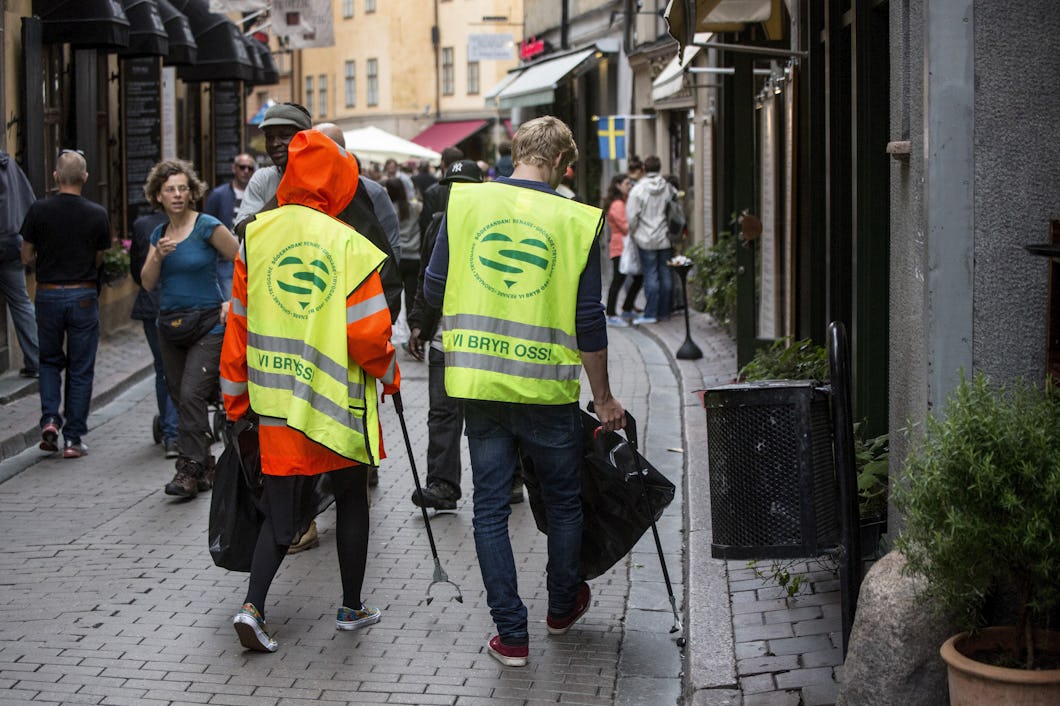 Två personer med gula säkerhetsvästar plockar skräp på en gata med gatsten i ett stadsområde medan andra går förbi.