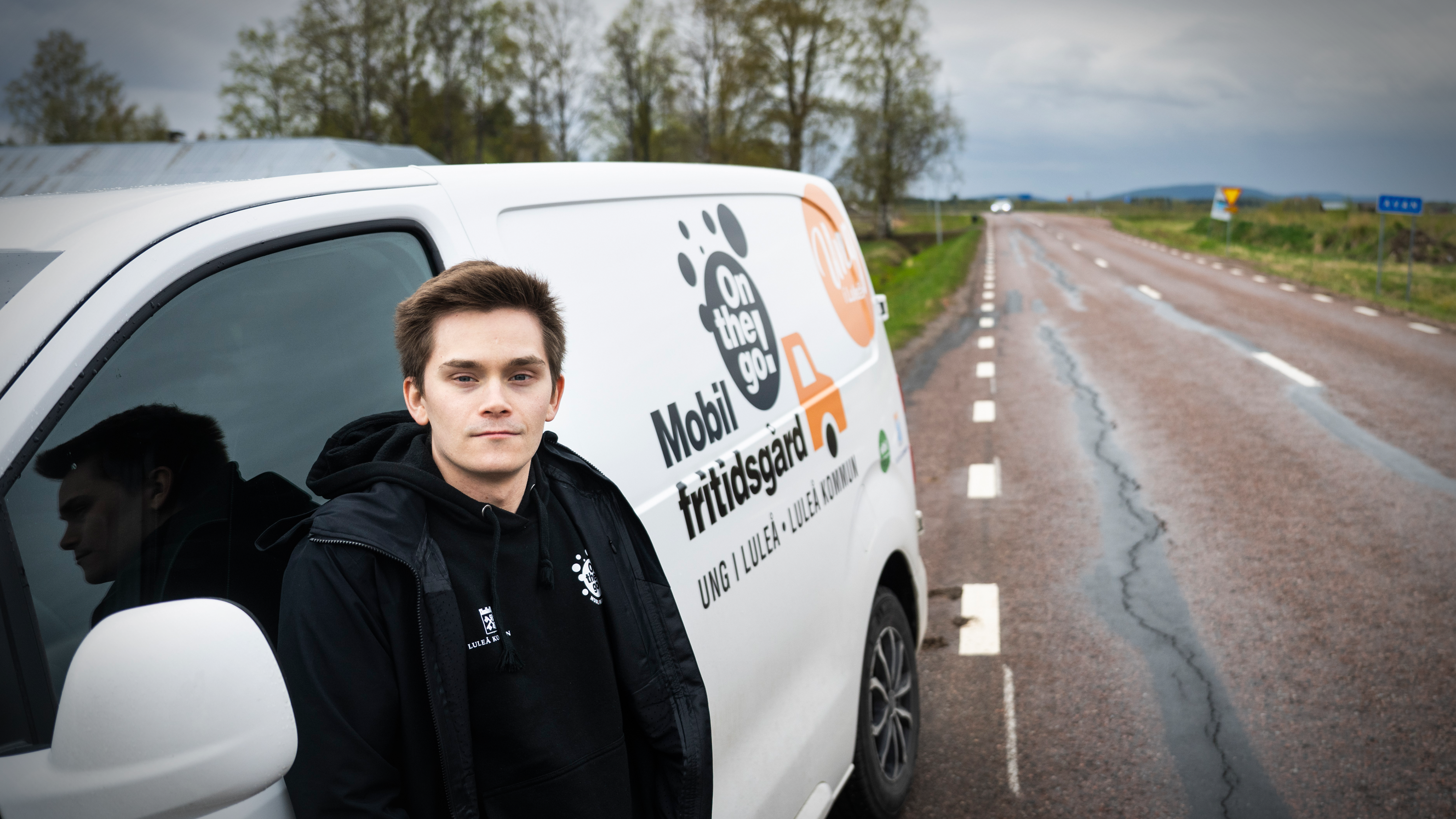 En ung man står bredvid en vit skåpbil med skylten "Mobil Fritidsgård". Skåpbilen är parkerad vid en landsväg under mulen himmel.