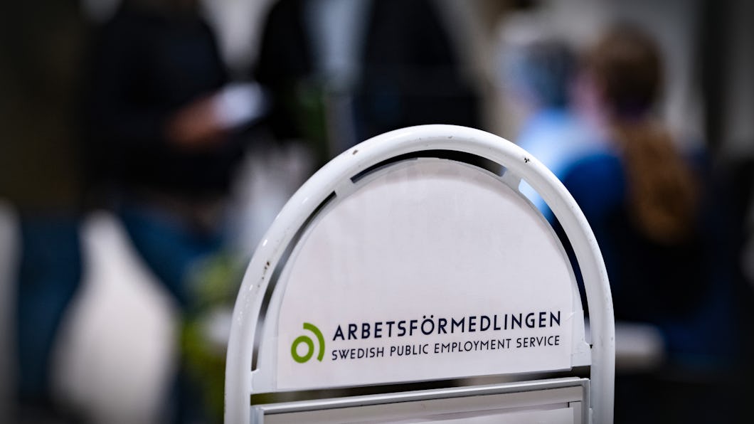 En skylt för Arbetsförmedlingen, den svenska myndigheten för arbetsförmedling, syns inomhus med personer i bakgrunden.
