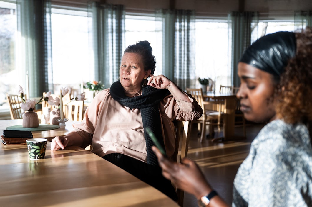 Marie Mattsson och Dure Dadacha sitter vid ett bord i ett rum med dagsljus. Marie tittar rakt fram, medan Dure tittar på sin mobiltelefon. På bordet finns koppar, anteckningsböcker och blommor.