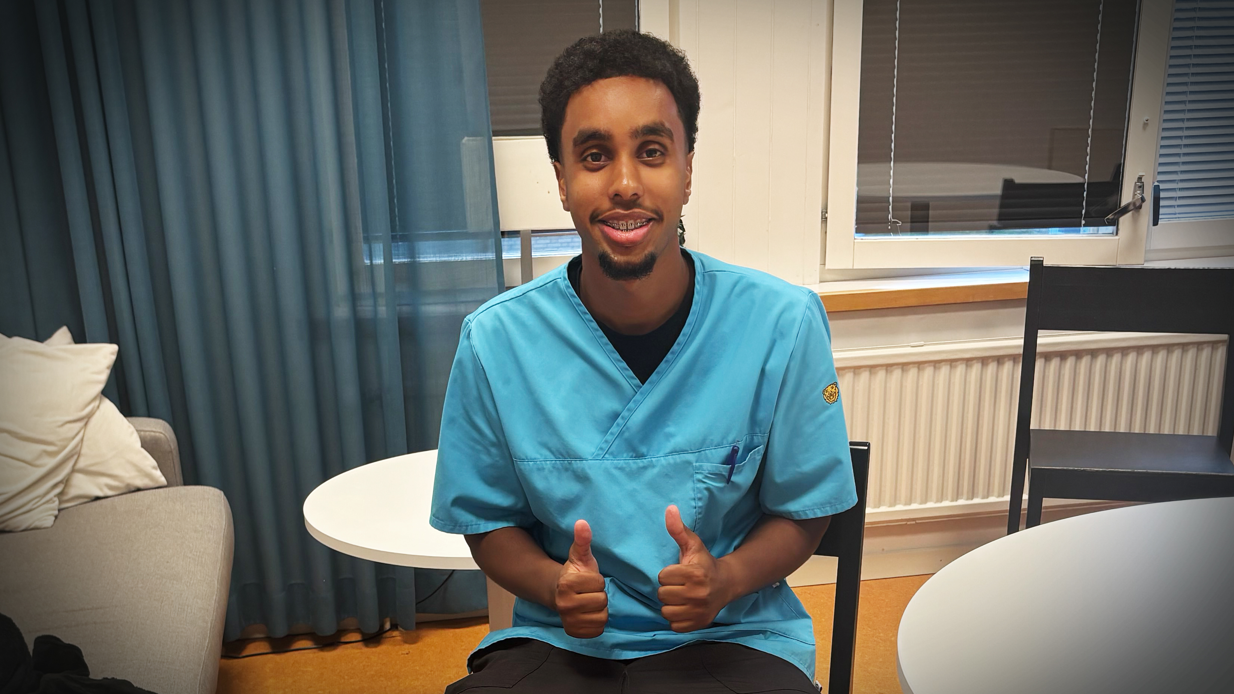 En man med blå sjukhuskläder sitter inomhus och ger två tummar upp. I bakgrunden finns stolar, ett bord och en soffa.