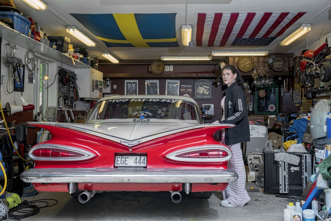 Pernilla Hendered står i ett garage bredvid en röd och vit äldre bil med texten "TOO FAT TO FLY" på bakrutan. Svenska och amerikanska flaggor hänger från taket.