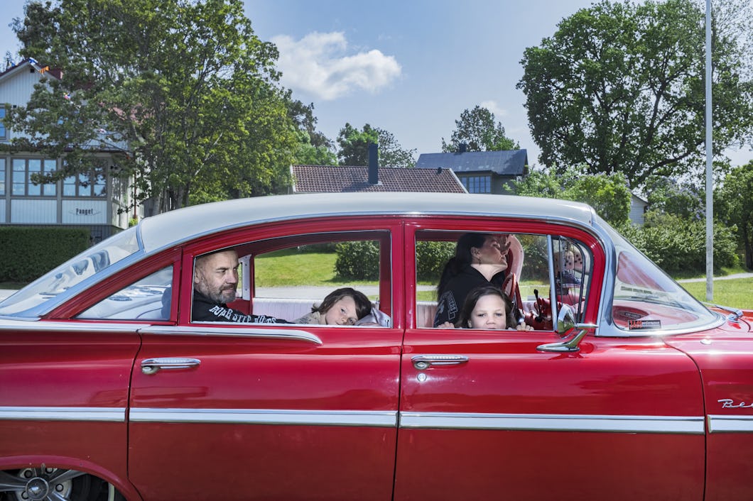 En familj sitter i en äldre röd bil som står parkerad på en gata. I bakgrunden finns träd och hus.