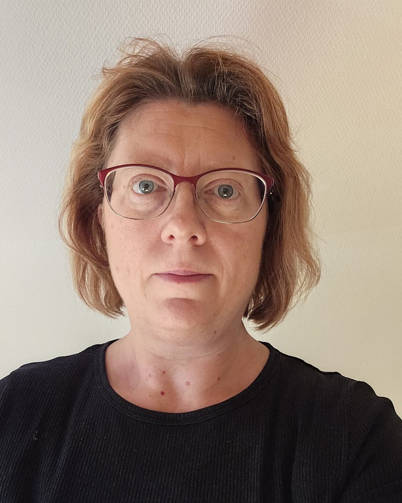En kvinna med röda glasögon och svart tröja tittar allvarligt in i kameran.