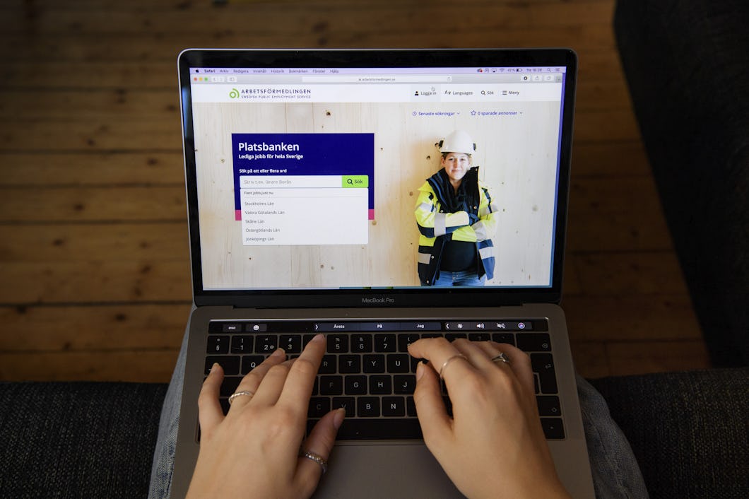 En person skriver på en MacBook som visar en jobbsökningssida med en bild av en byggarbetare med skyddsväst och hjälm på skärmen.