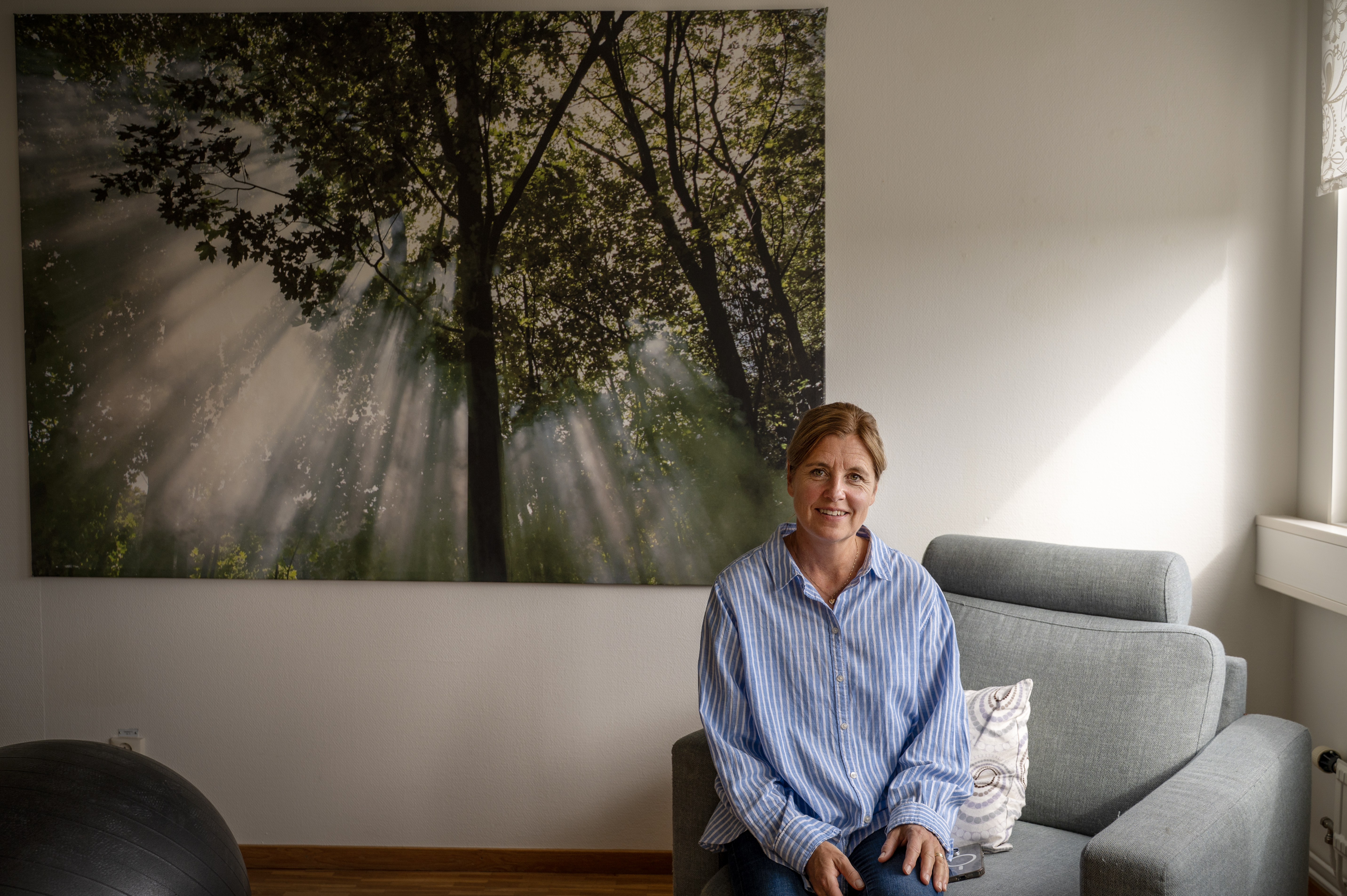 En kvinna sitter i en grå fåtölj i ett rum, med en stor tavla med träd bakom sig.