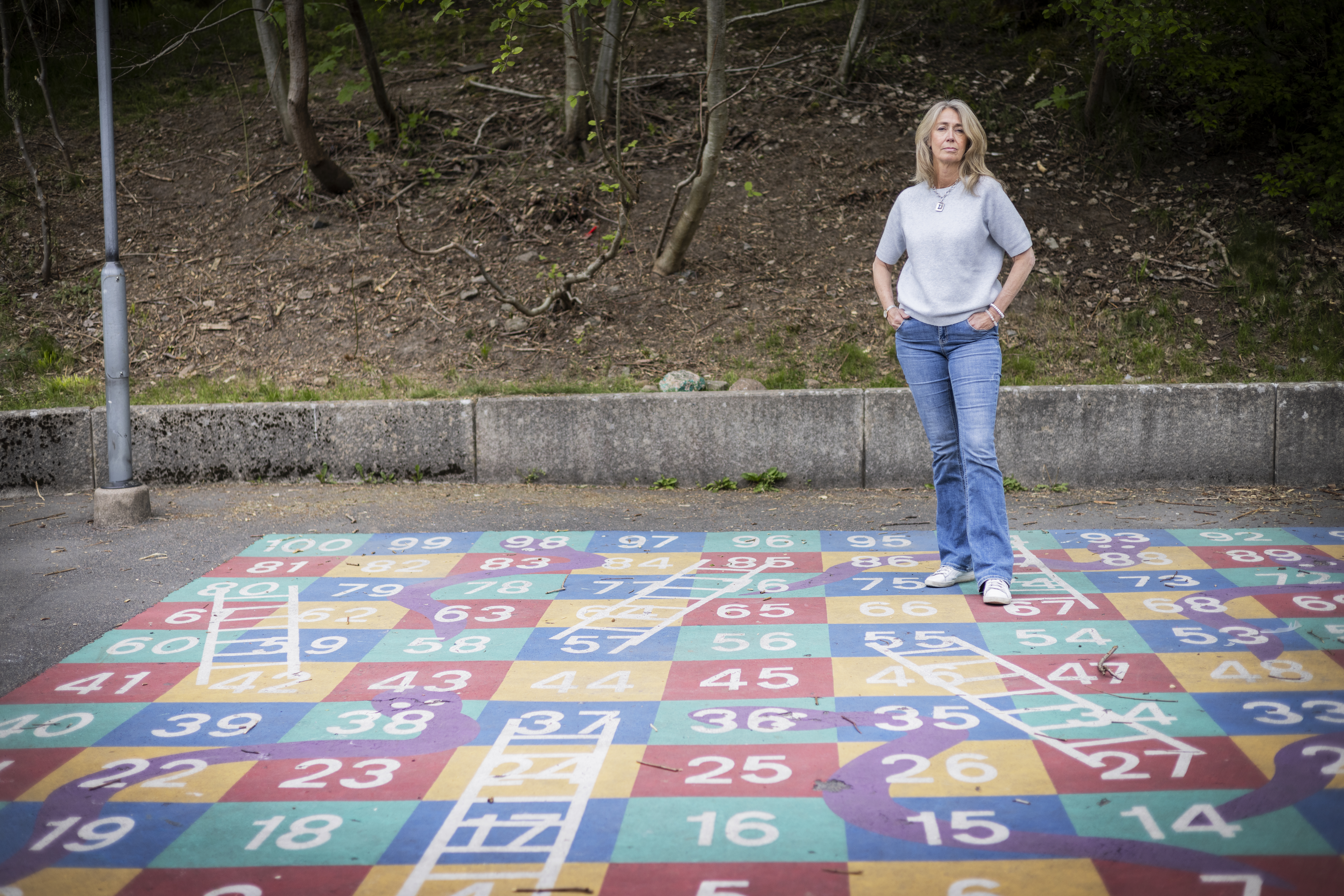 Birgitta Larsson står på en lekplats med rutor som har siffror och målade ormar och stegar, med ett skogsområde i bakgrunden.