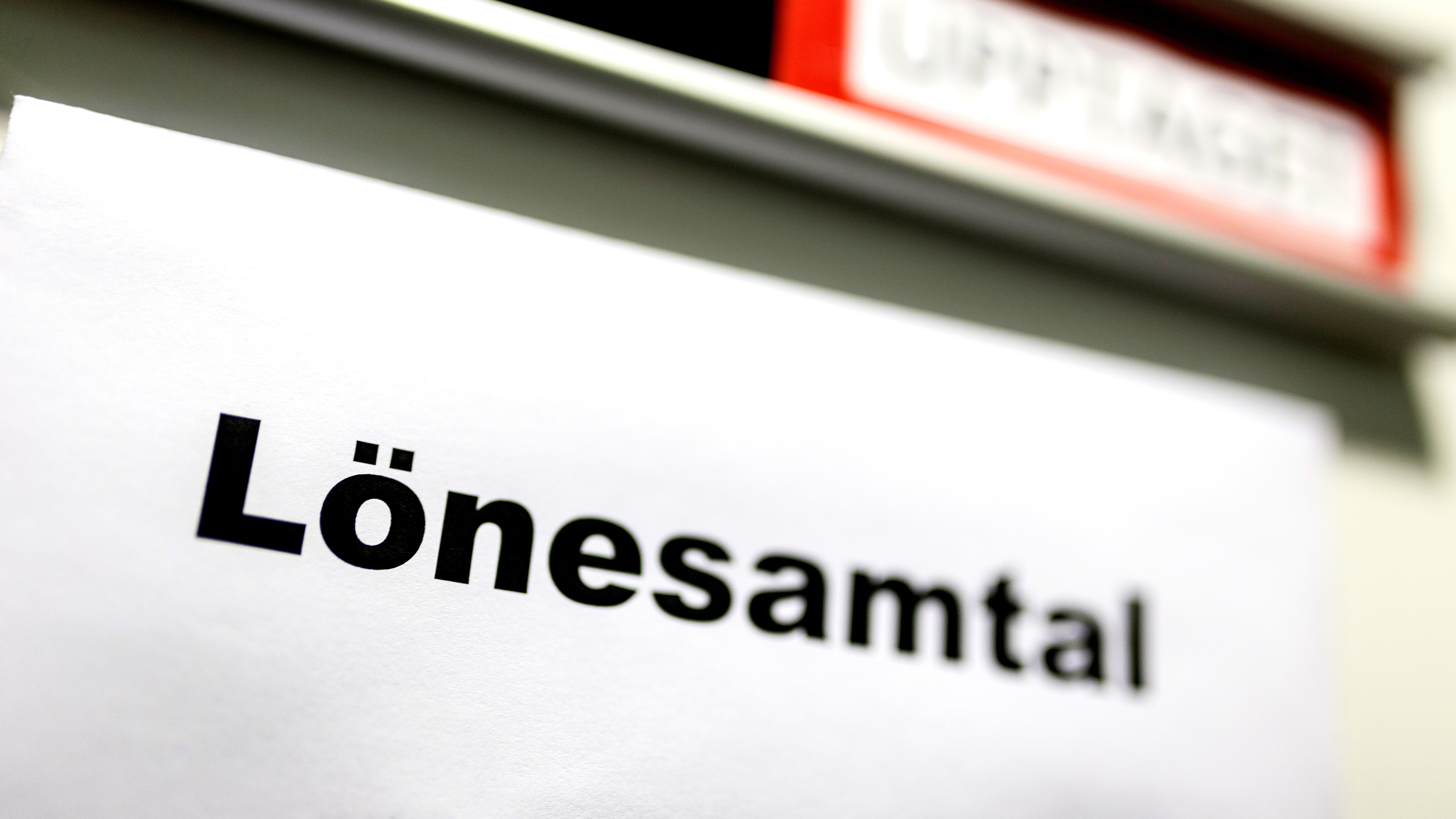 Ett papper med ordet "Lönesamtal" på svenska sitter fast på en yta. Ovanför syns en del av en röd och vit skylt.