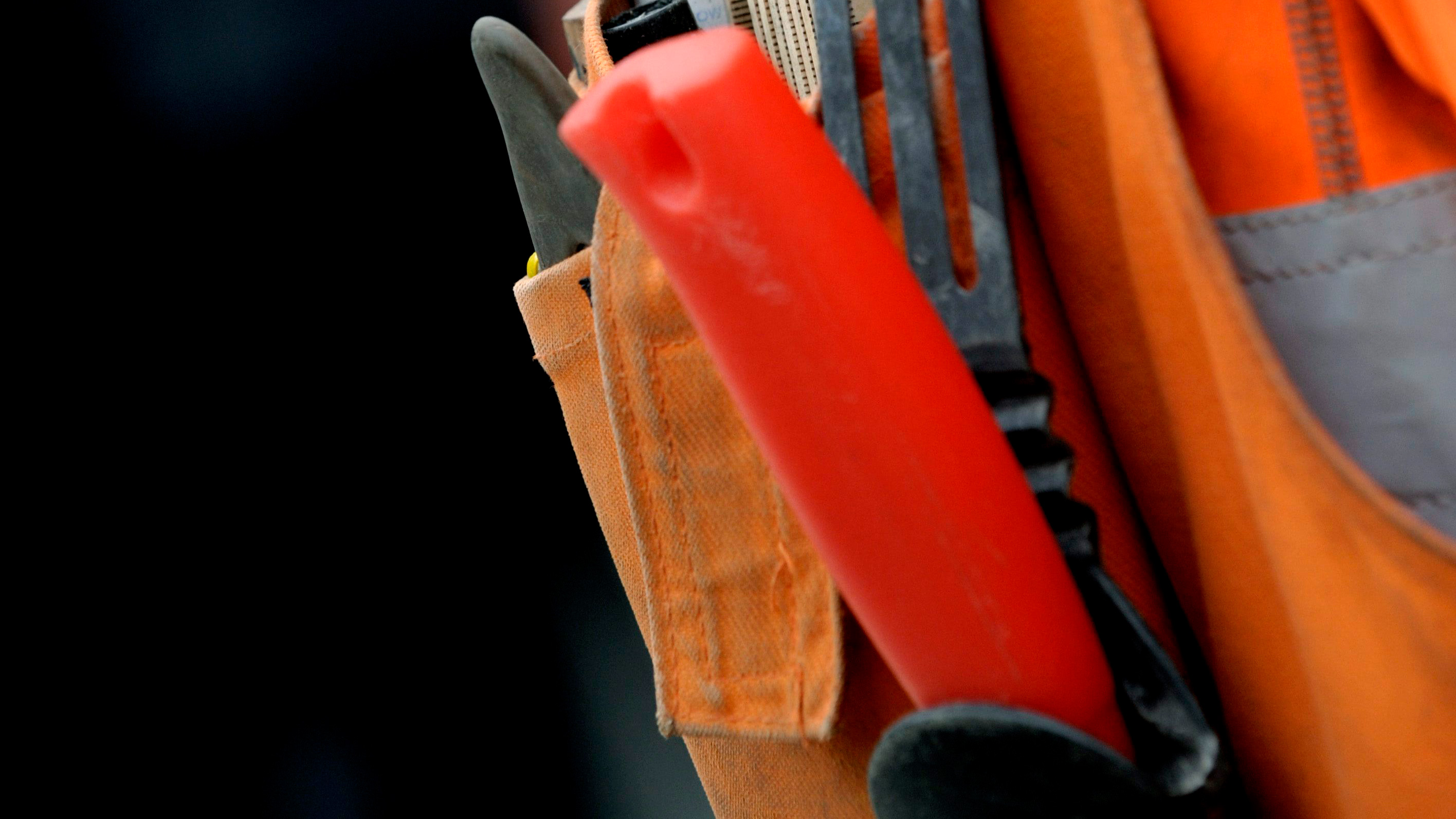 Närbild på en verktygsficka på en orange arbetsväst med ett rött knivhandtag och andra svarta verktyg.