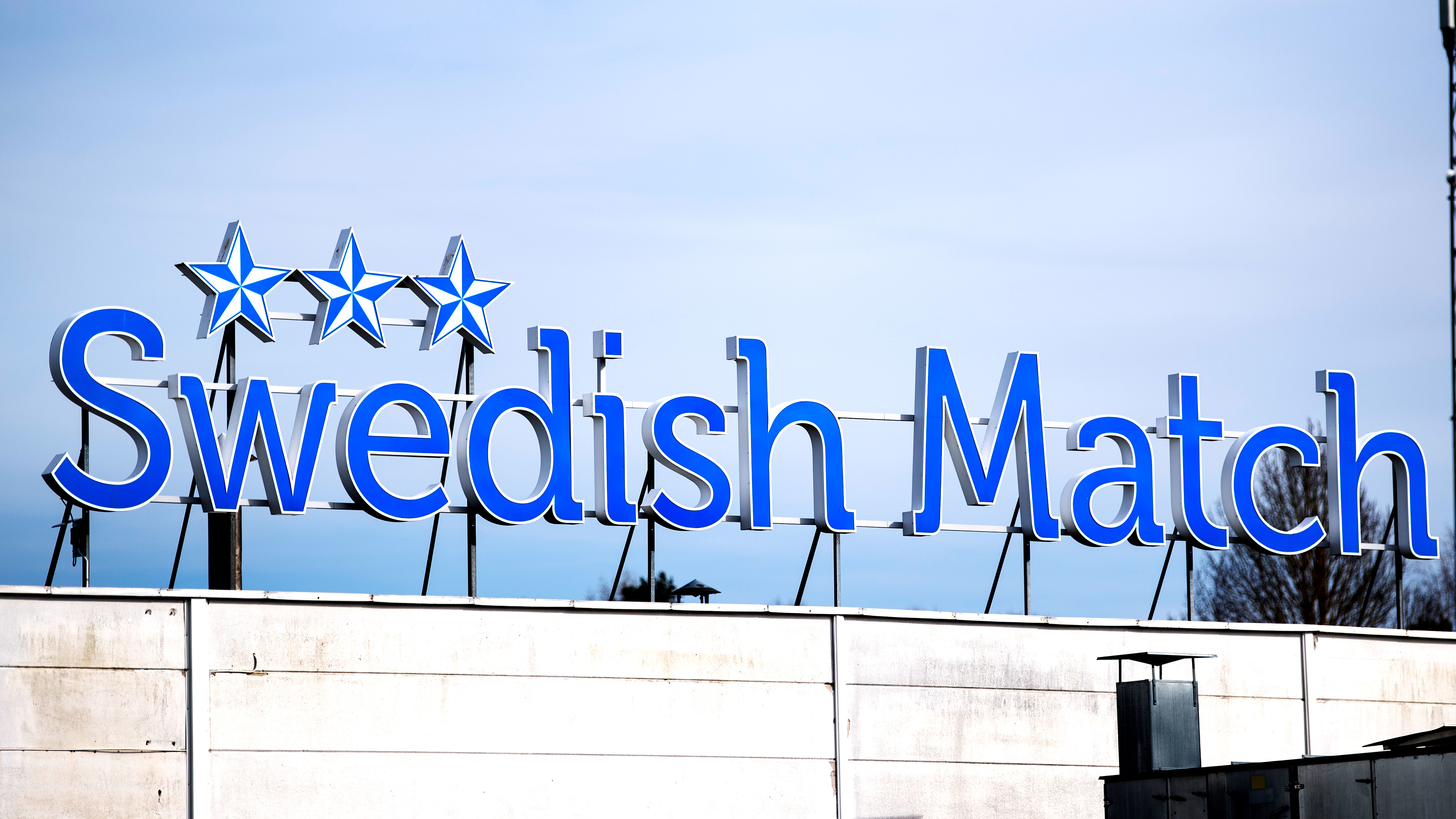 Blå "Swedish Match"-skylt med tre stjärnor syns ovanpå en vit byggnad mot en klar himmel.