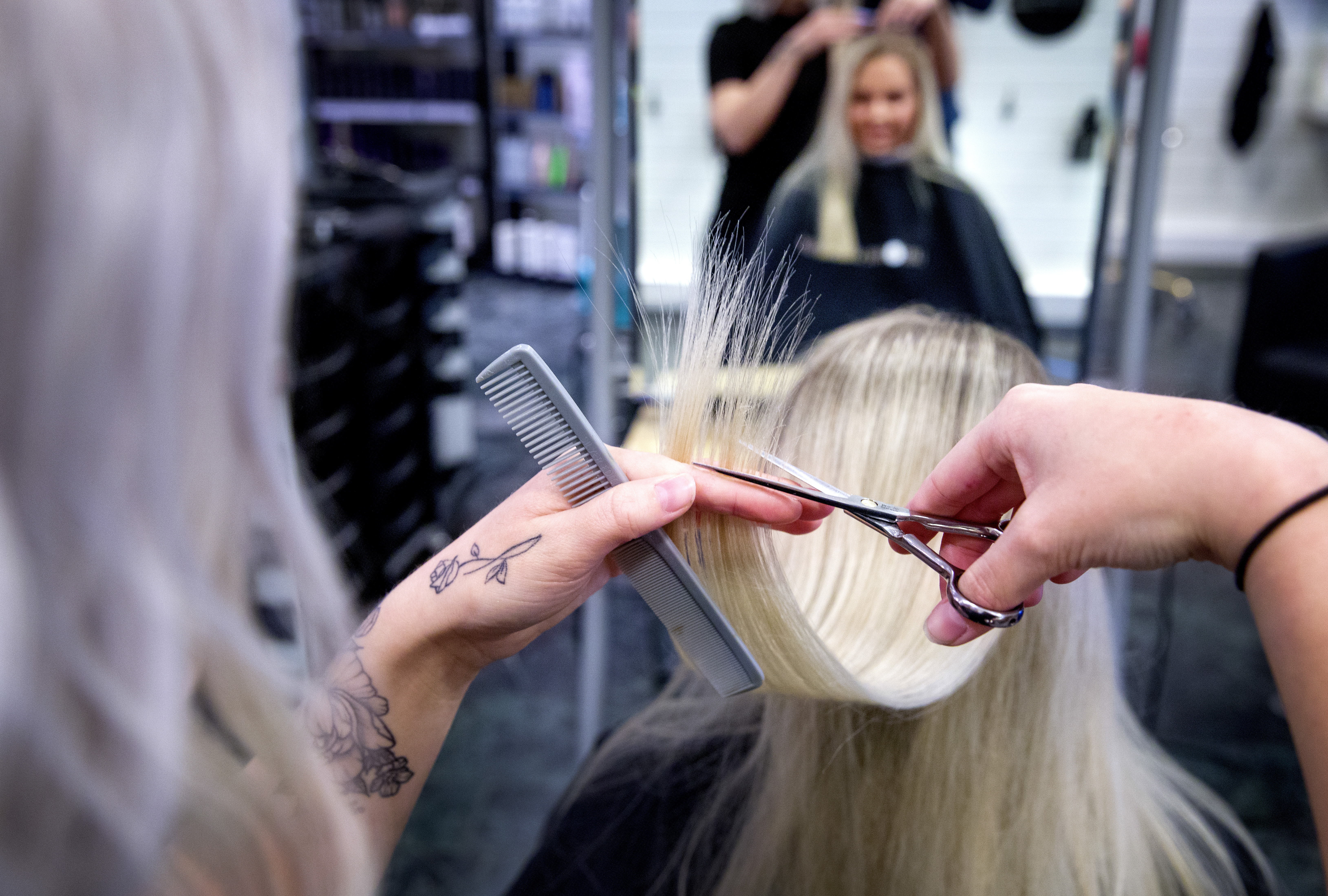 En frisör klipper en kunds blonda hår med sax och kom i en salong, medan kunden ser på sin spegelbild.