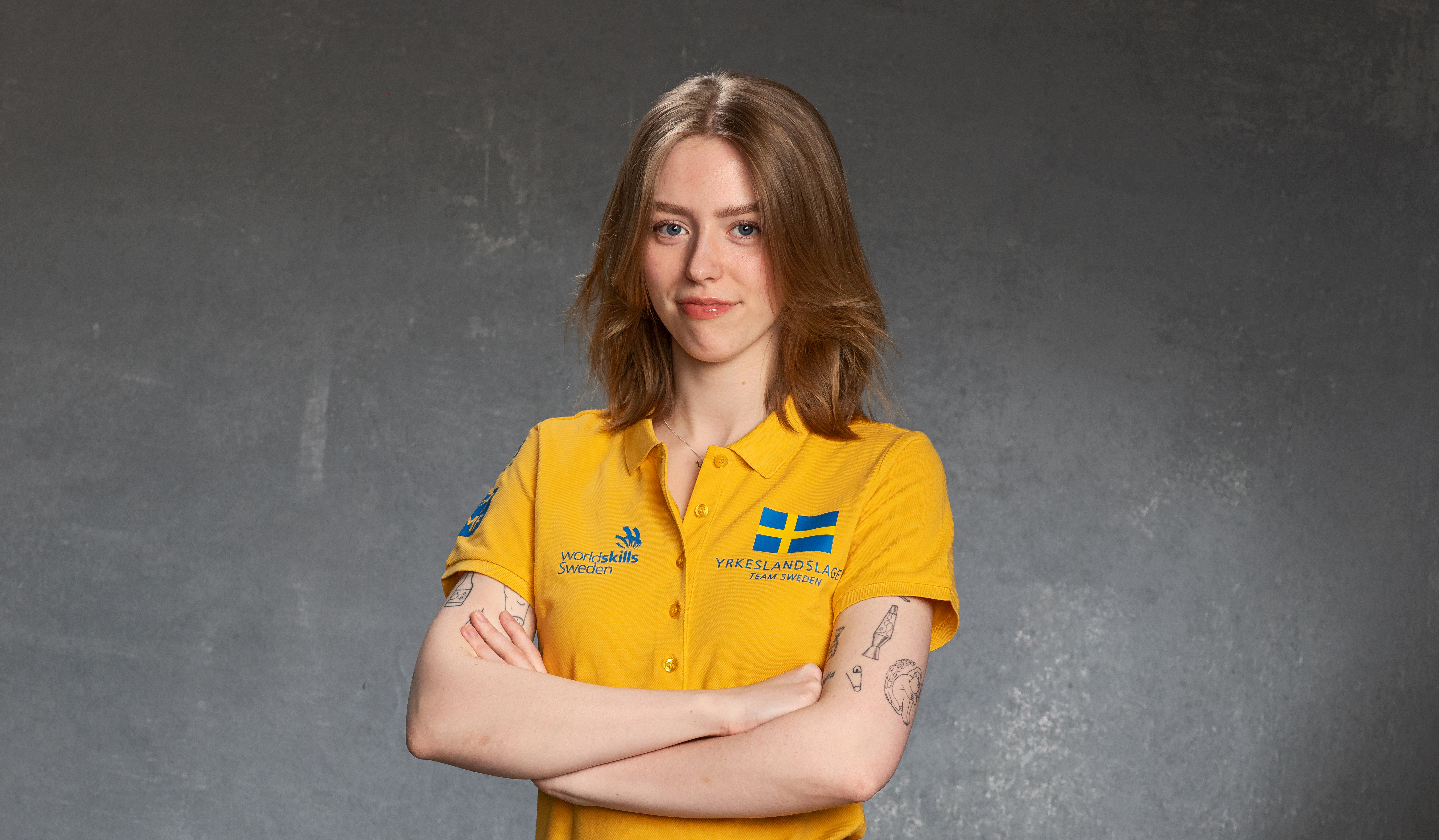 Kvinna i gul t-shirt står med armarna i kors och tittar i kameran.