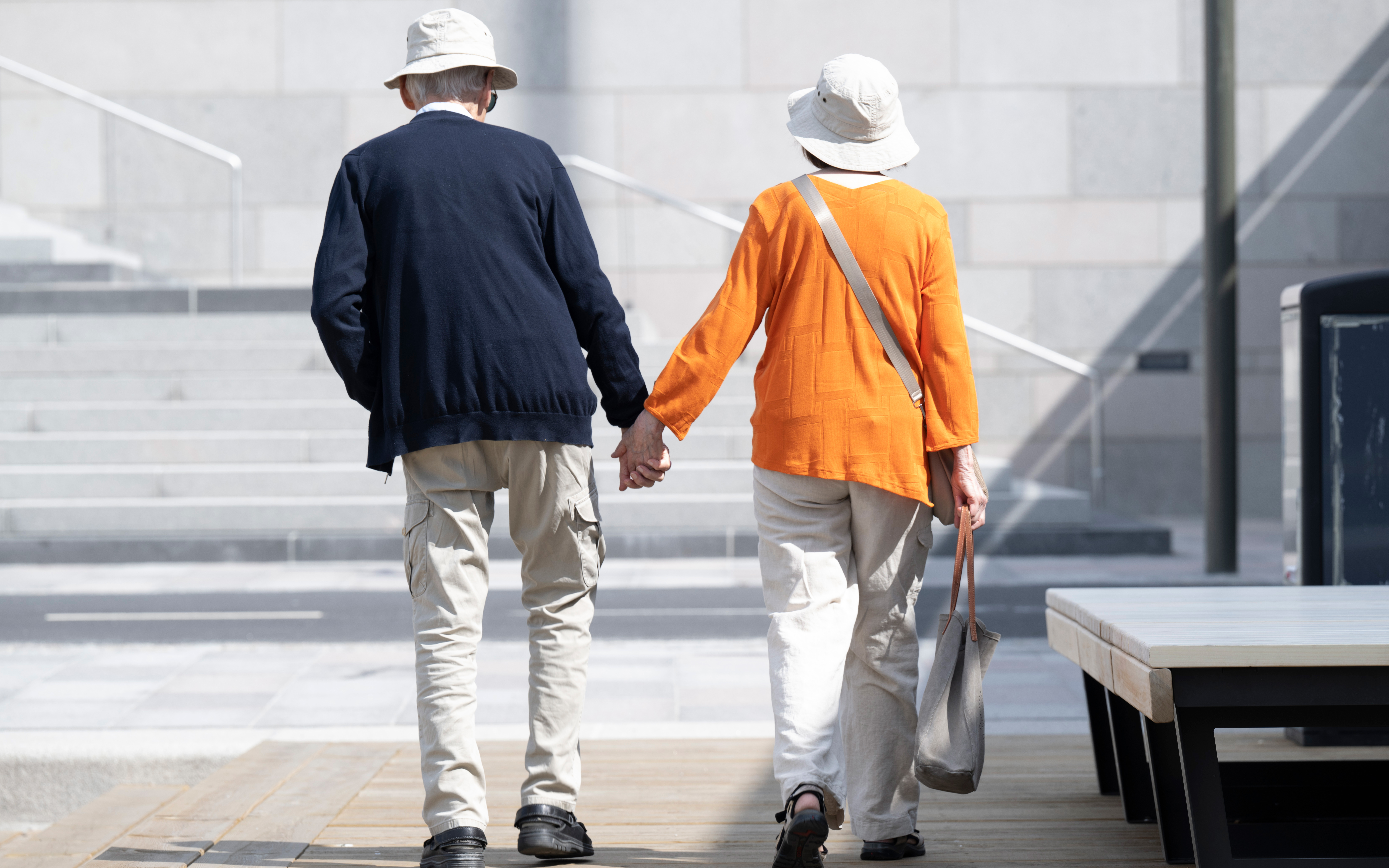 Ett äldre par med hattar och lediga kläder går hand i hand utomhus nära trappor och sittplatser.