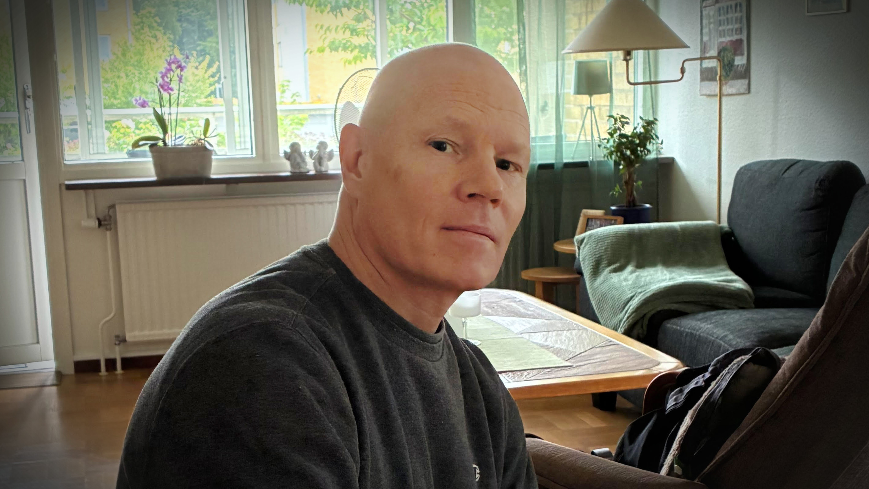 Krister Maltby med en mörk tröja sitter i ett vardagsrum med ett bord, växter och ljus från stora fönster i bakgrunden.