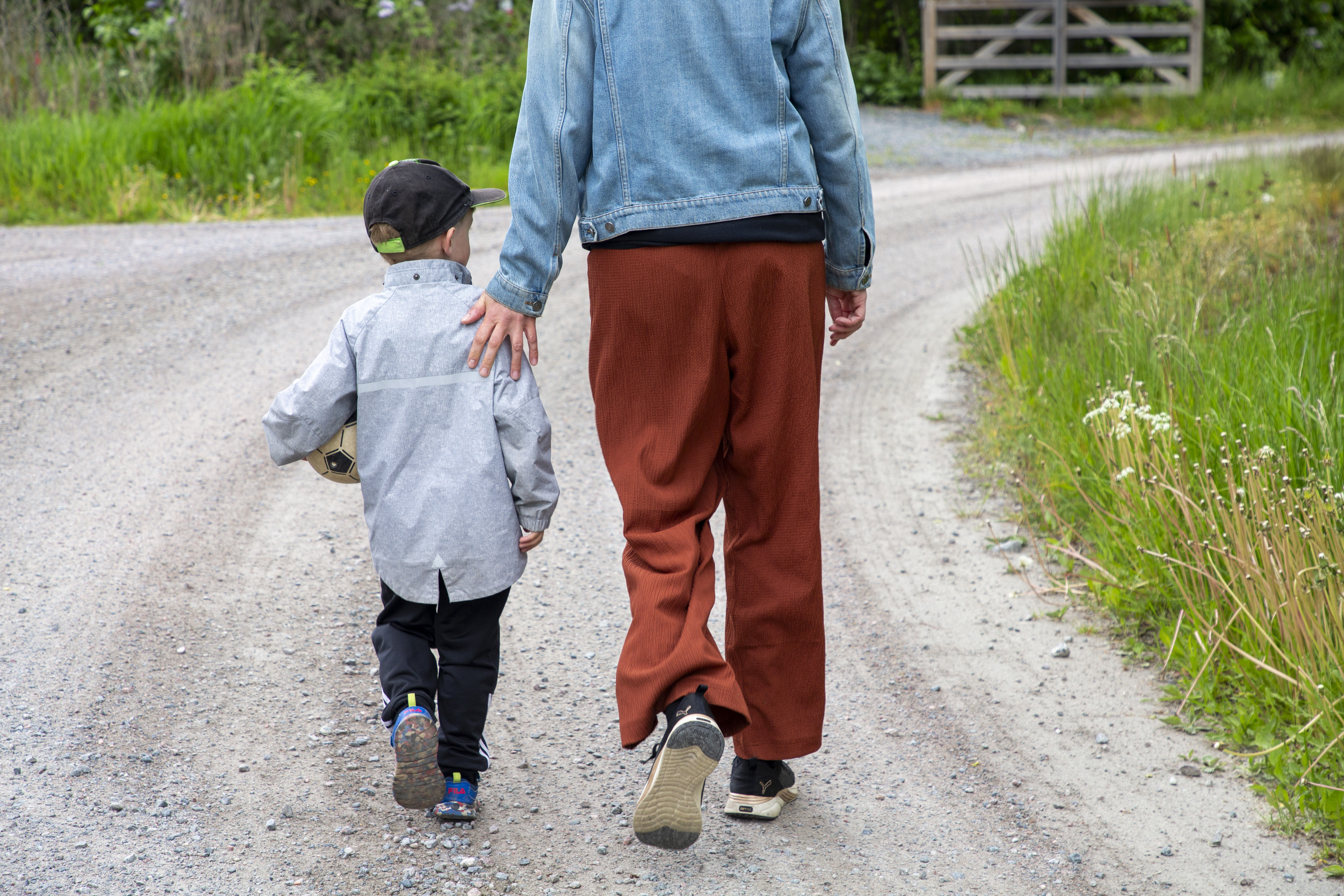 En vuxen och ett barn går hand i hand på en grusväg. Barnet bär en boll under ena armen. De omges av växter.
