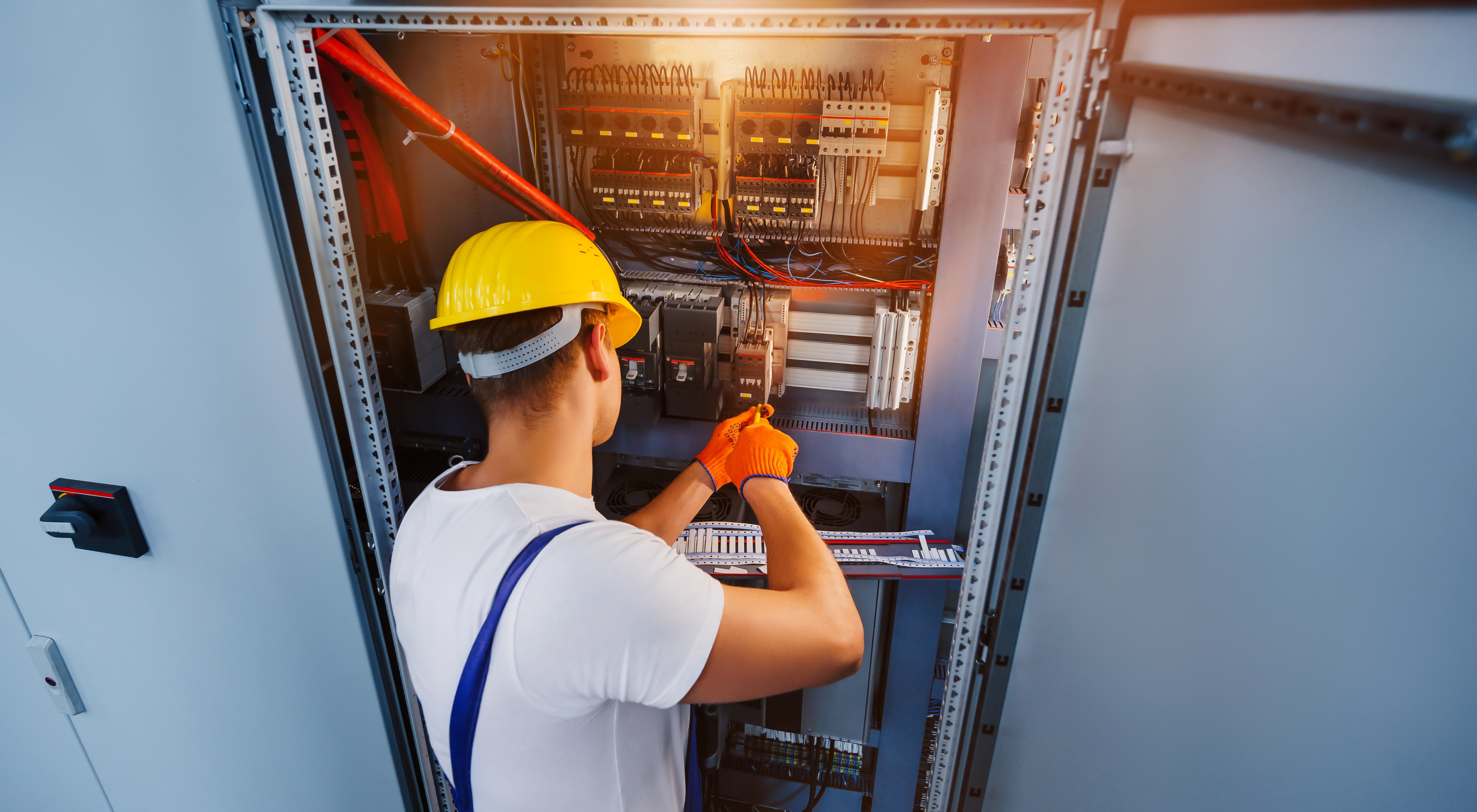 En elektriker med gul hjälm och orange handskar arbetar med kablage i ett öppet elskåp.