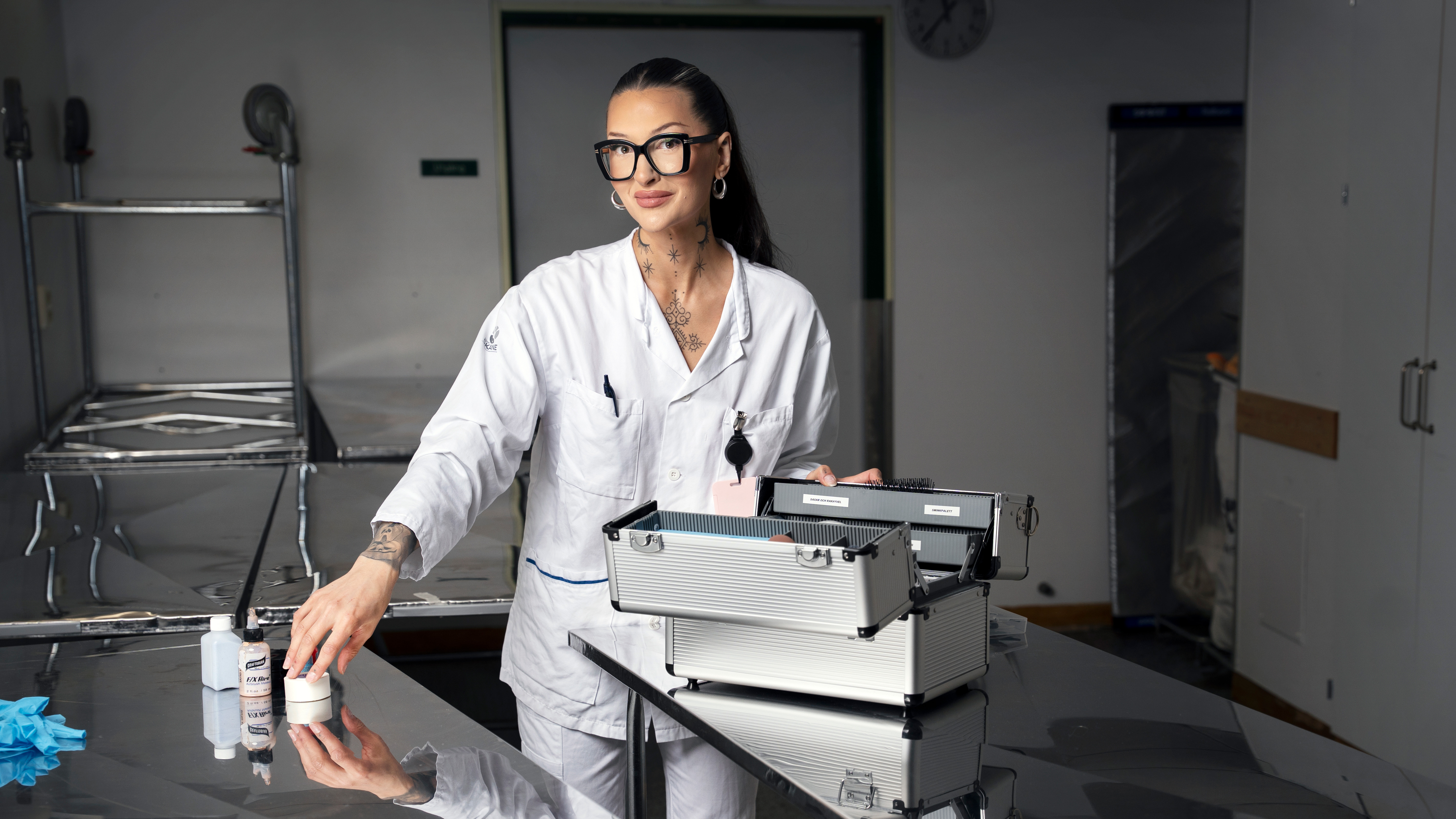 Angelika Andersson i vit labbrock står vid ett bord av rostfritt stål med medicinska artiklar och metallådor i en klinisk miljö.