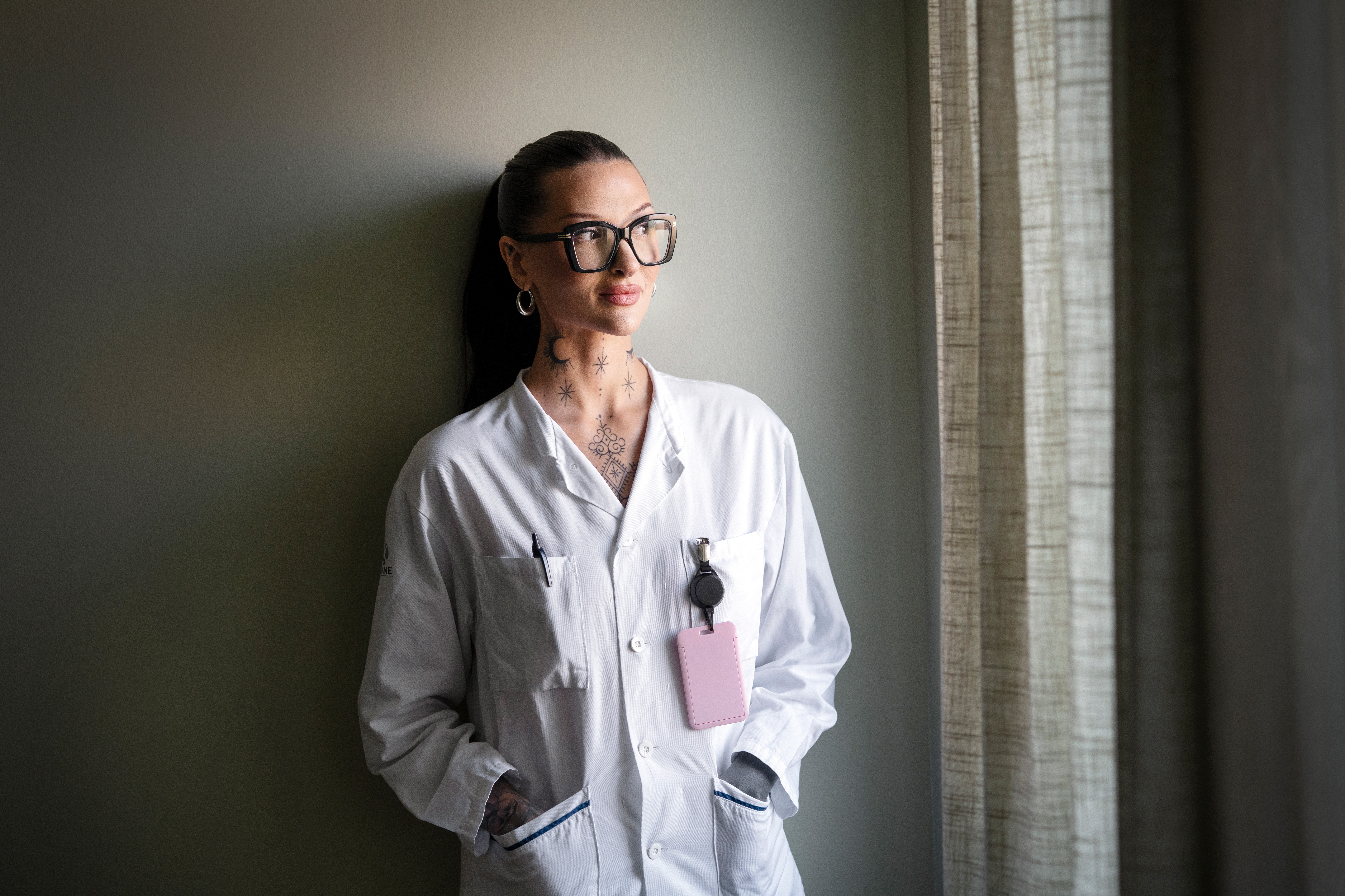 Person i vit laboratorierock med ID-bricka står vid ett fönster och tittar ut. Personen har glasögon, tatueringar och långt bakåtdraget hår.