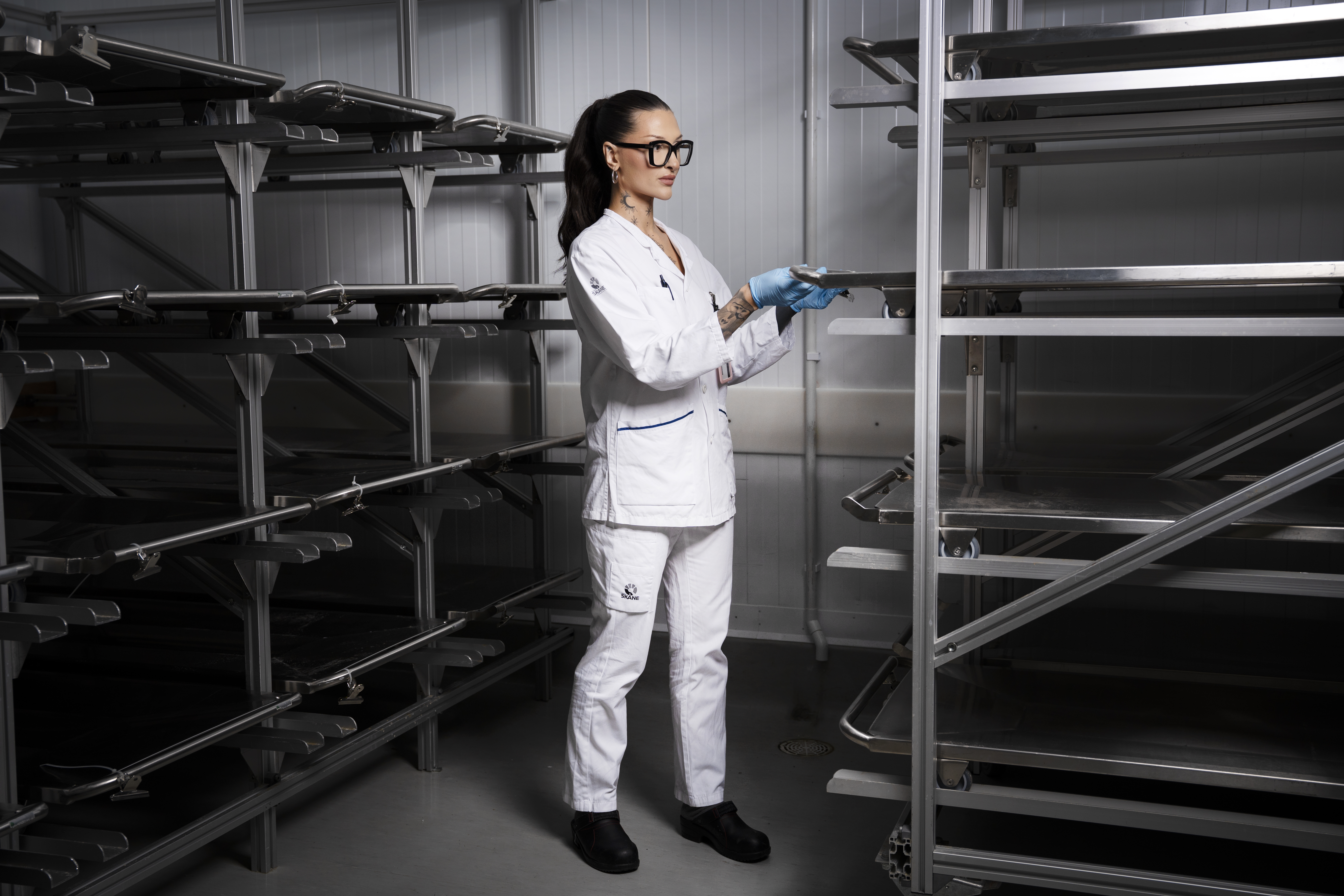 Angelika Andersson med vit labbrock, handskar och glasögon står bredvid tomma metallställ i en industriell miljö.