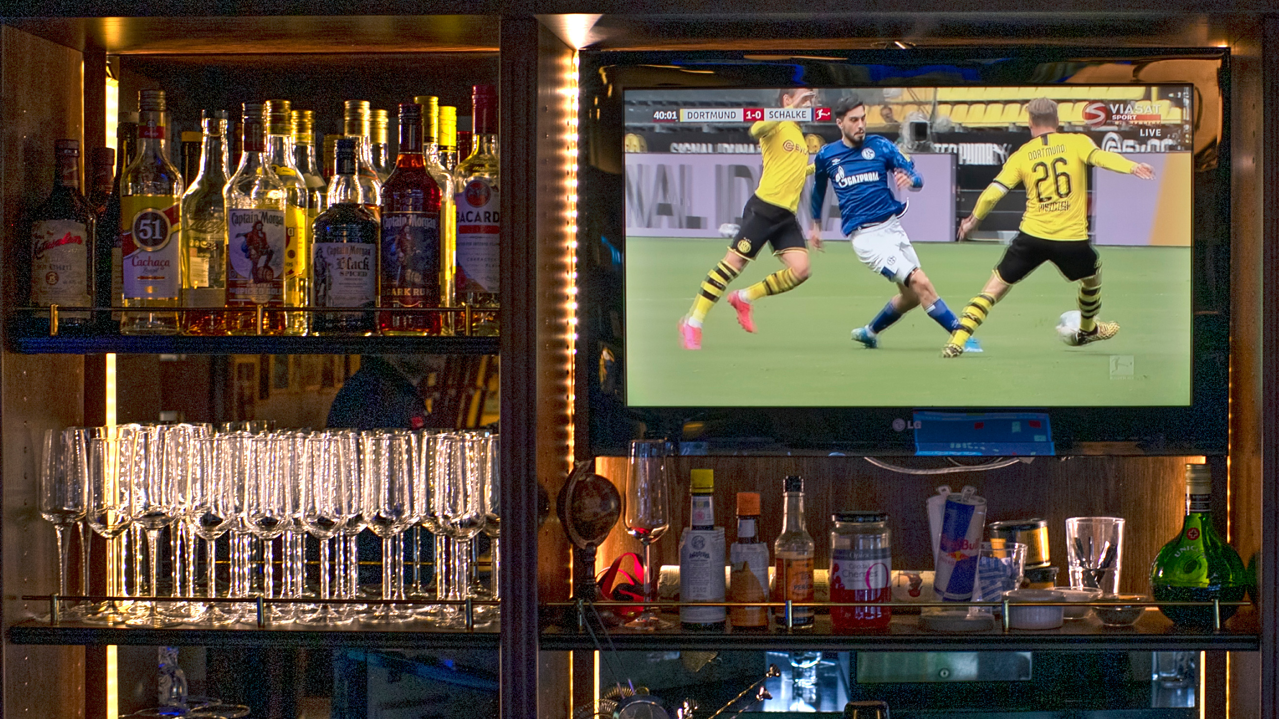 Flaskor och glas står framme i en bar, med en tv ovanför som visar en fotbollsmatch mellan lag i gula och blå tröjor.