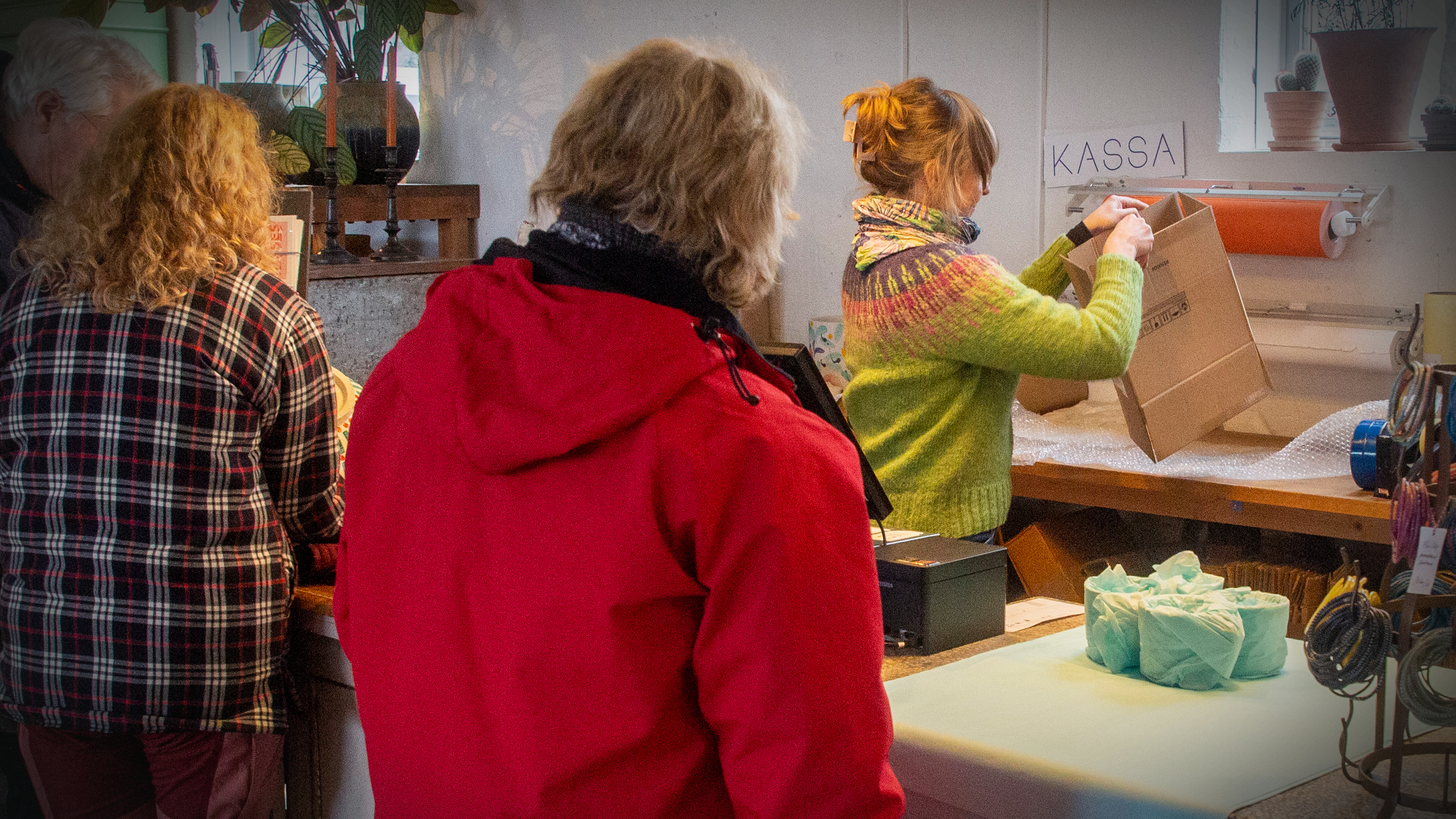 Flera personer står i kö vid en disk med skylten ”kassa” medan en anställd packar saker i en kartong.