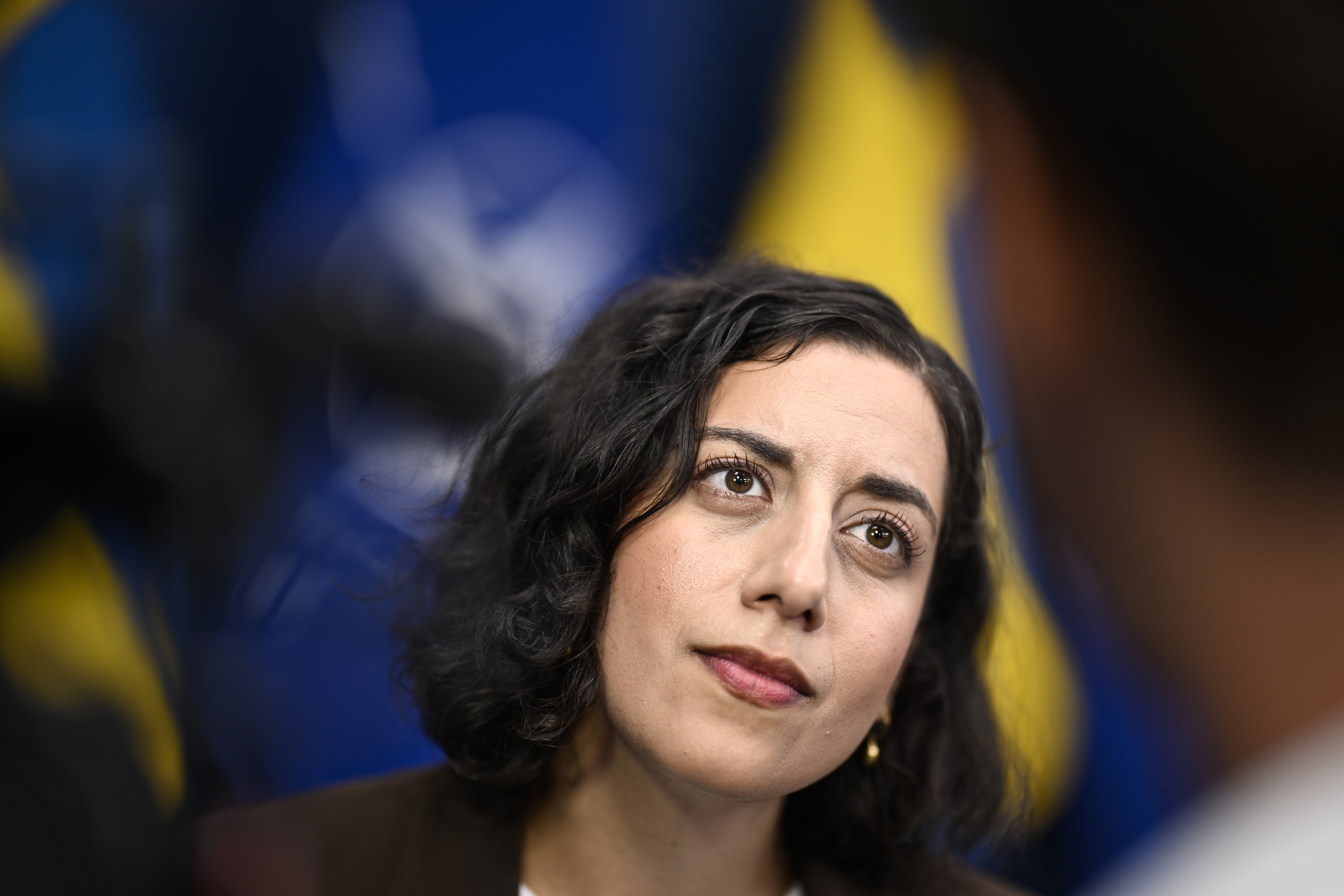 Simona Mohamsson med mörkt, lockigt hår tittar på någon utanför kameran. I bakgrunden finns blå och gula flaggor.