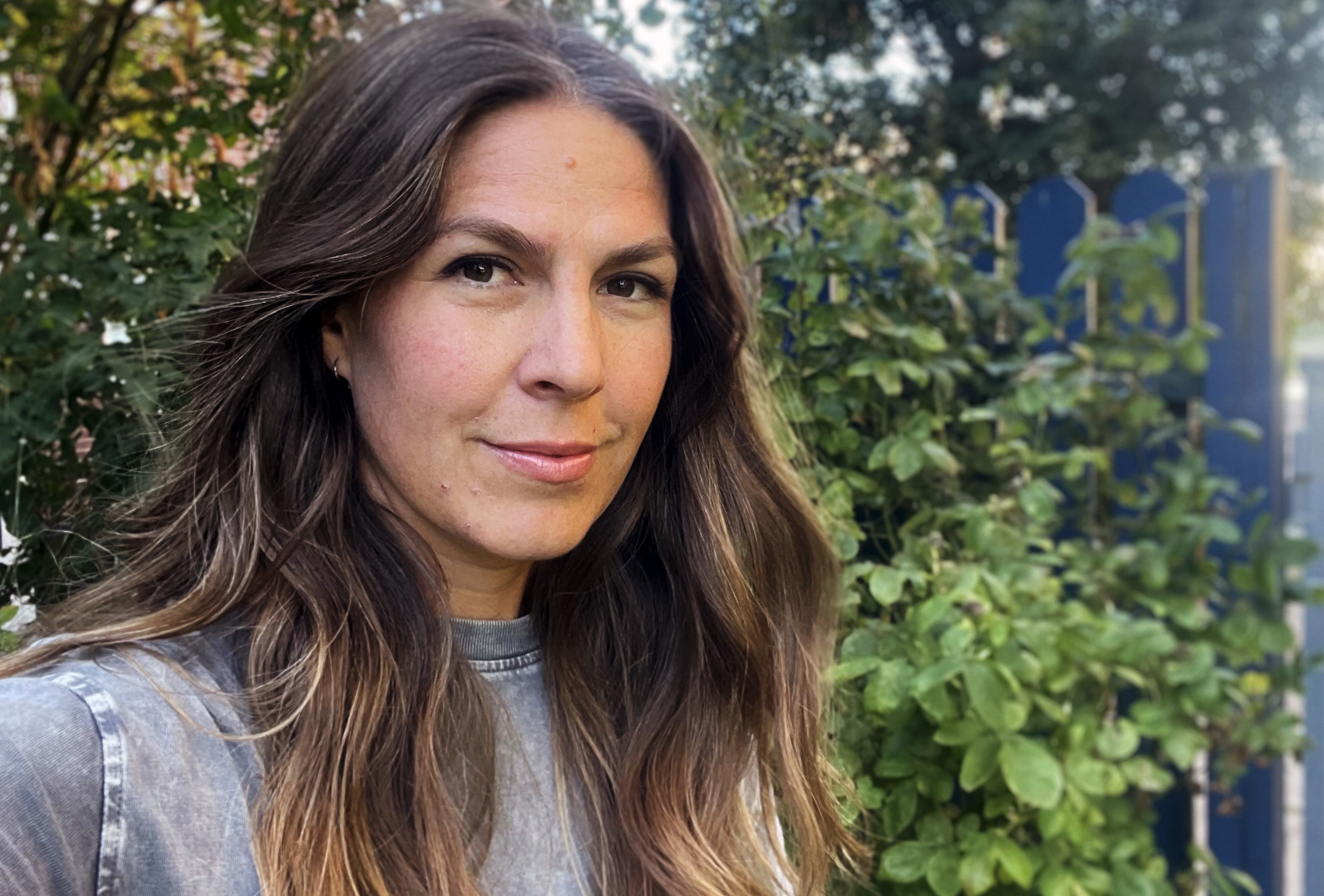 Anna Wingård med mörkt långt hår och grå tröja står framför träd och ett blått staket.