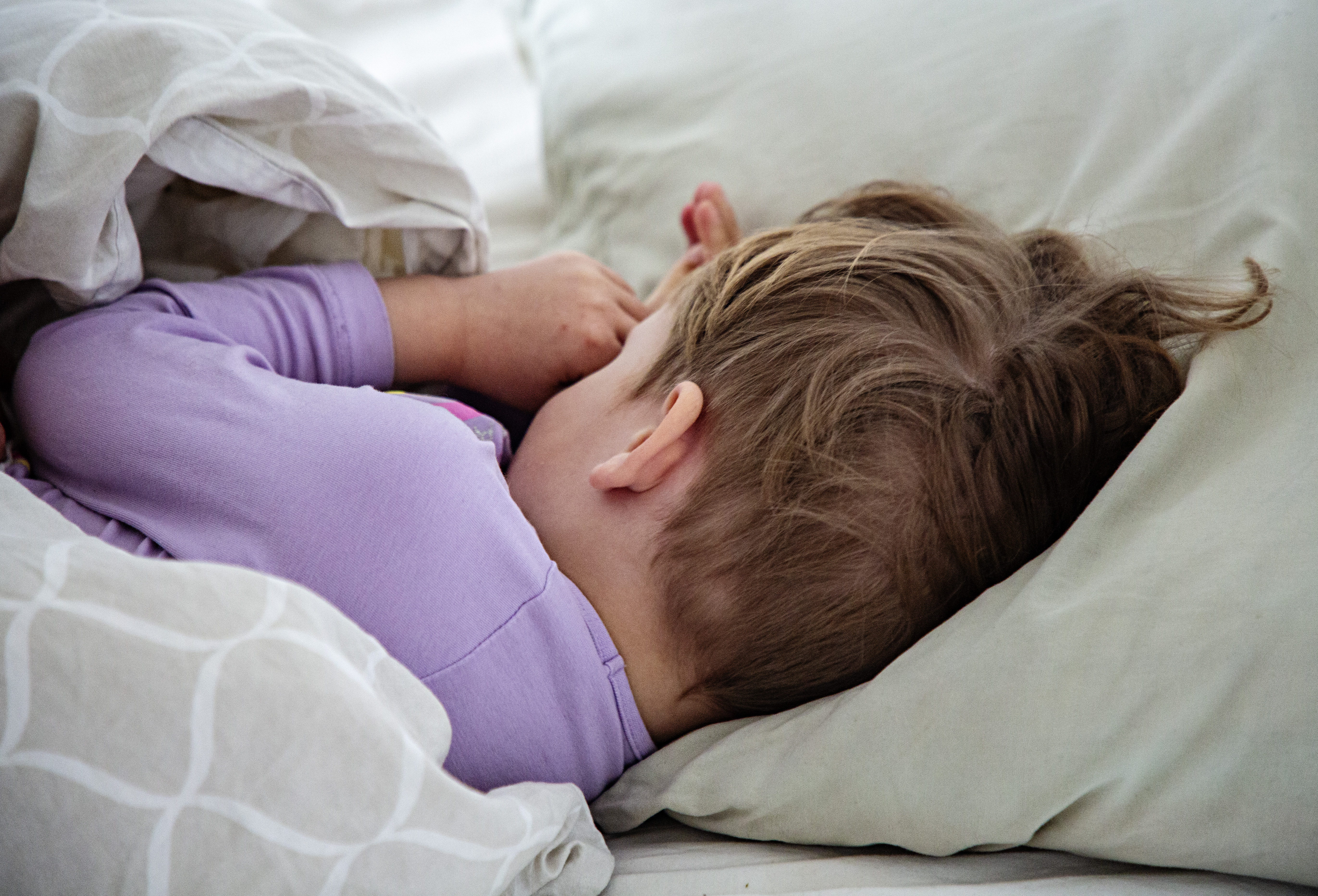 Ett barn med lila tröja ligger på sidan i sängen, sover med huvudet på en kudde och täcke delvis över sig.