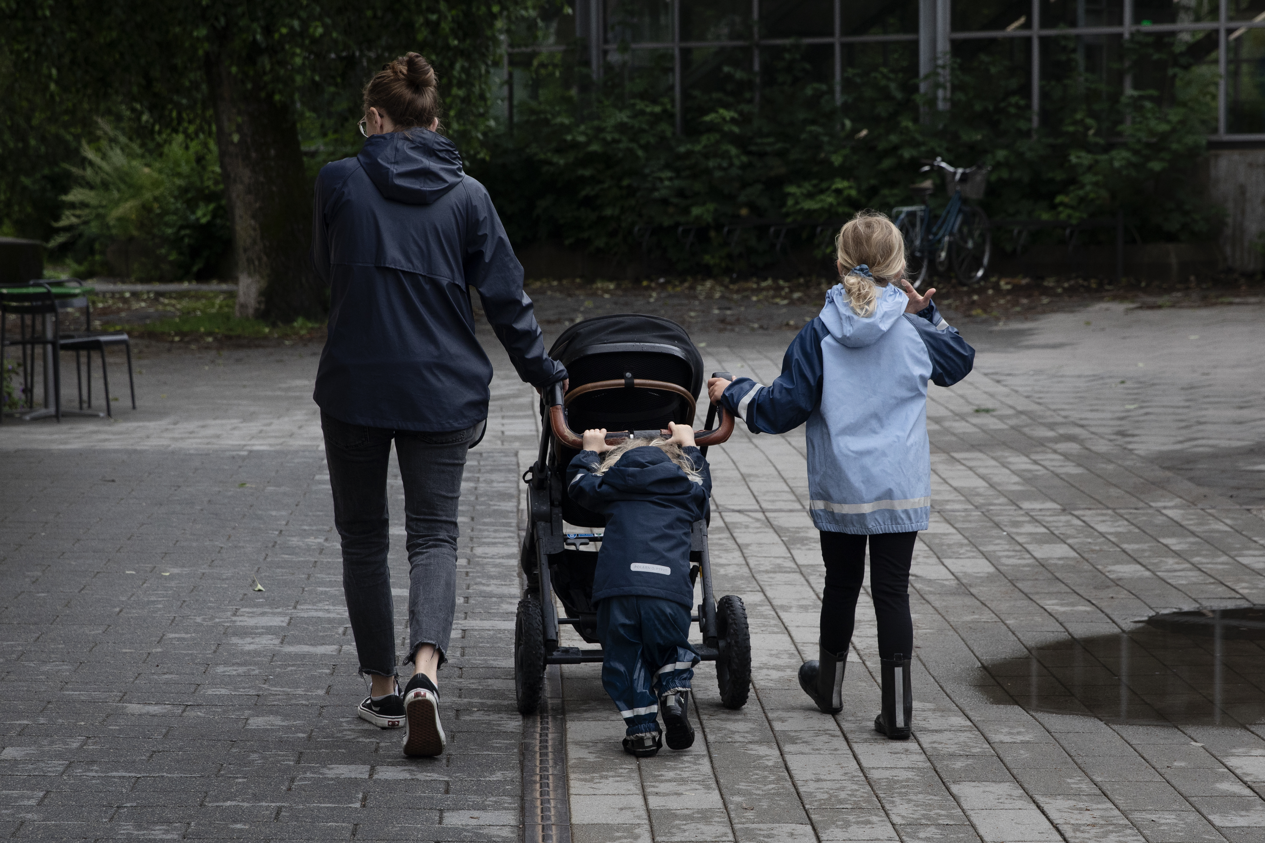 En vuxen och två barn, varav ett skjuter en barnvagn, går tillsammans på en asfalterad väg nära växter och en byggnad.