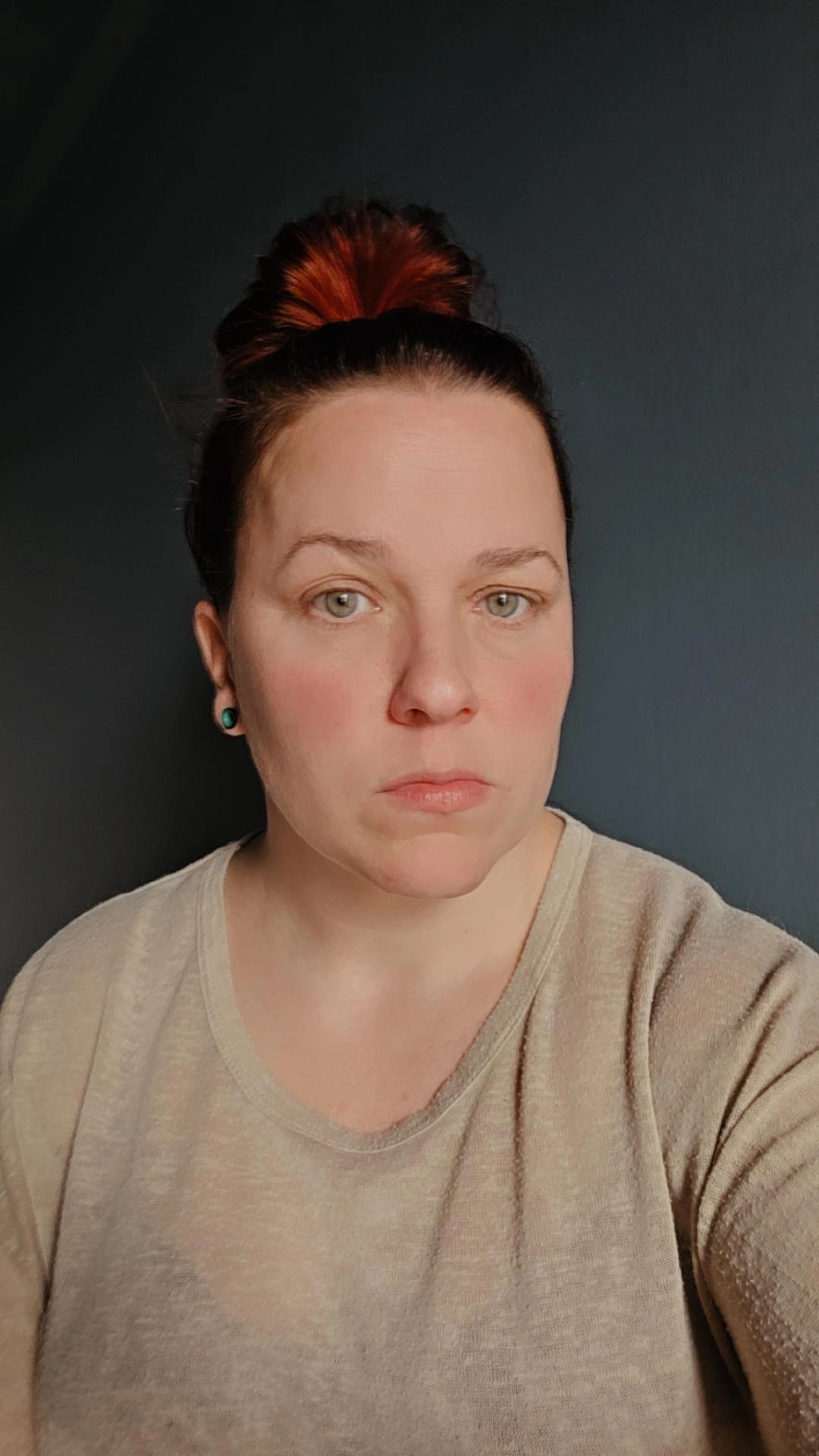 En kvinna med ljus hud och rödbrunt hår i en knut bär en beige topp och tittar rakt in i kameran mot en mörk bakgrund.