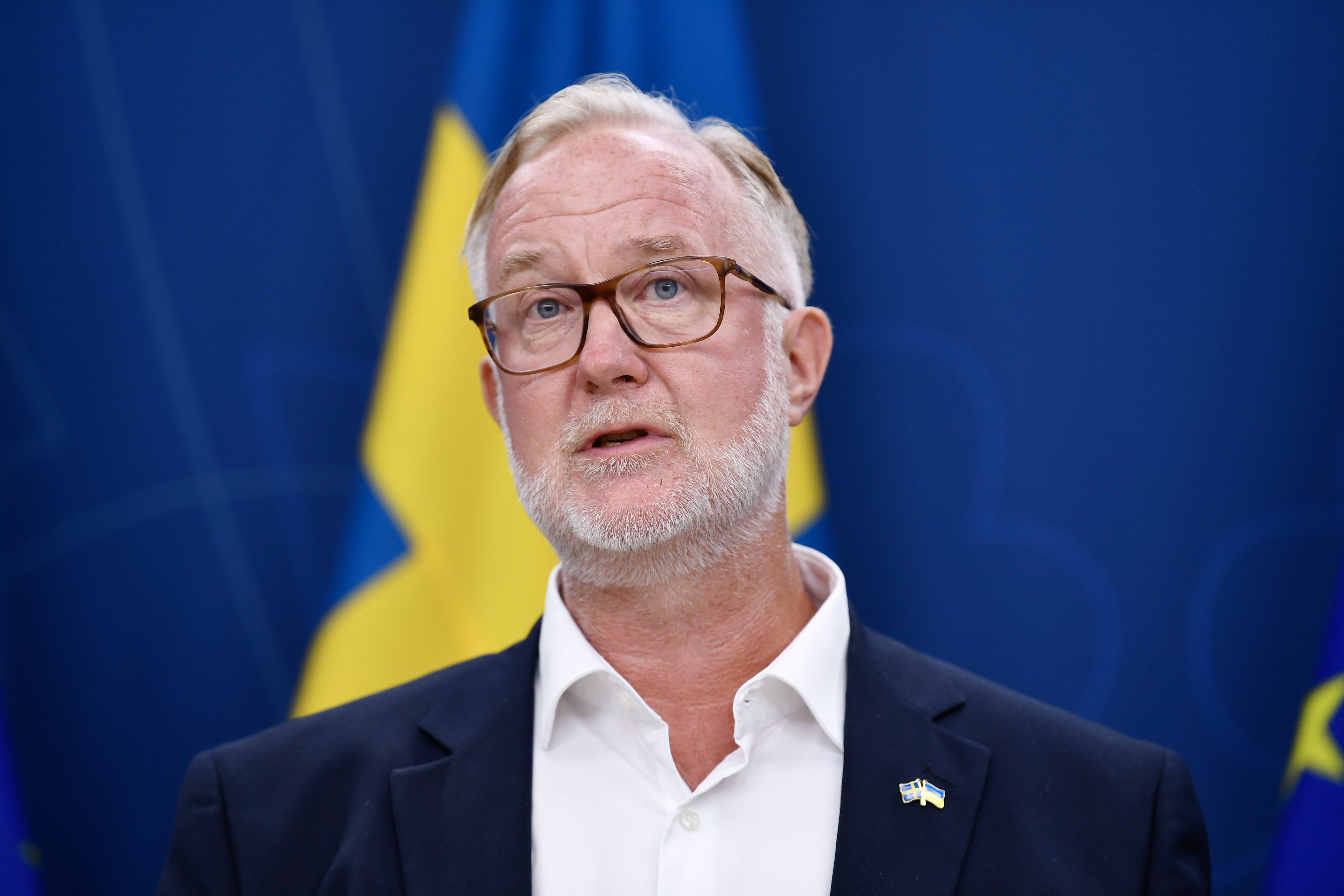 Johan Pehrson med glasögon och vitt skägg talar framför blå och gula flaggor, iförd kavaj med en liten flaggknapp på slaget.