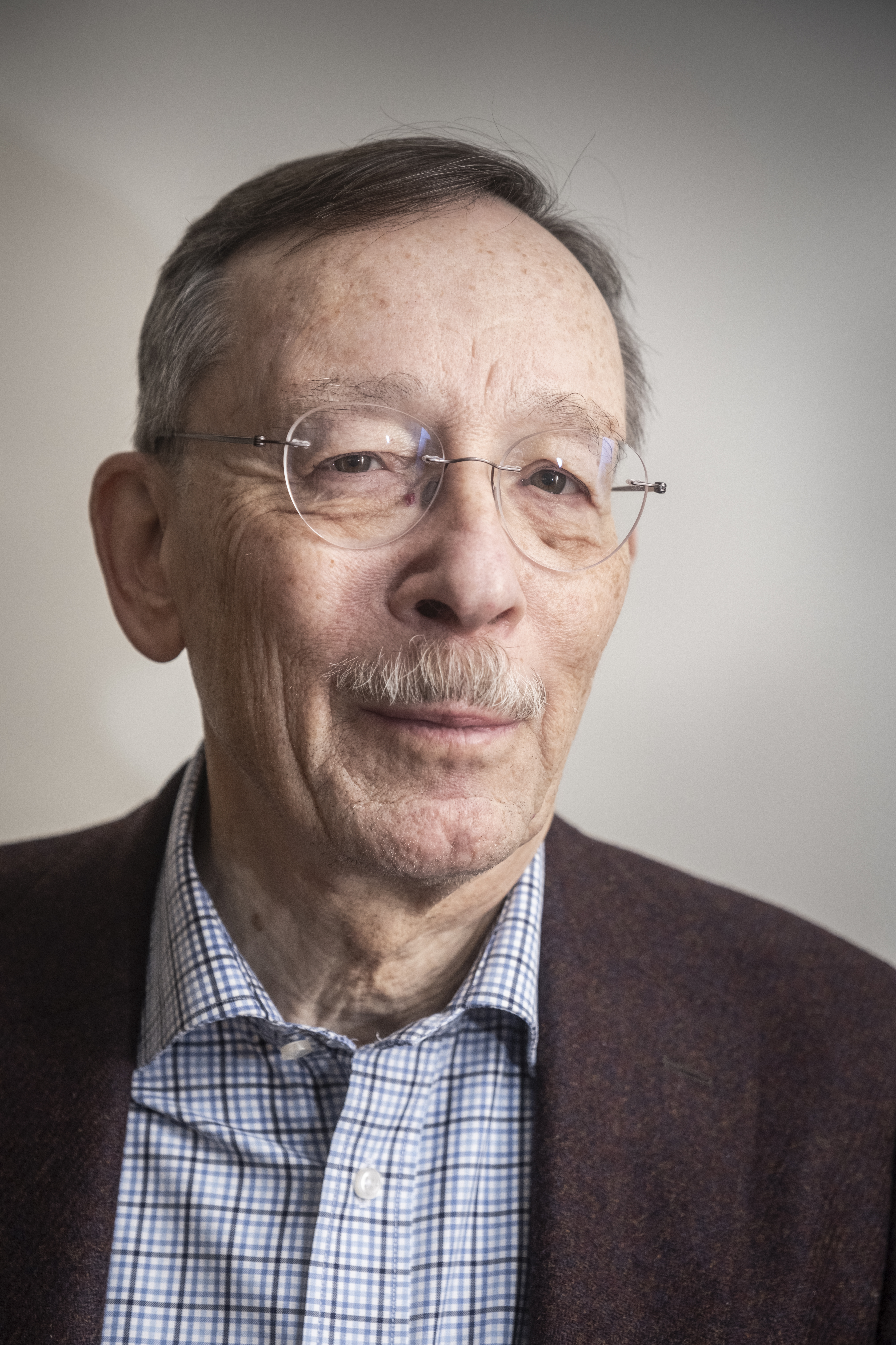 Lars Calmfors med glasögon, mustasch och kort grått hår har på sig en rutig skjorta och mörk jacka mot en enkel bakgrund.