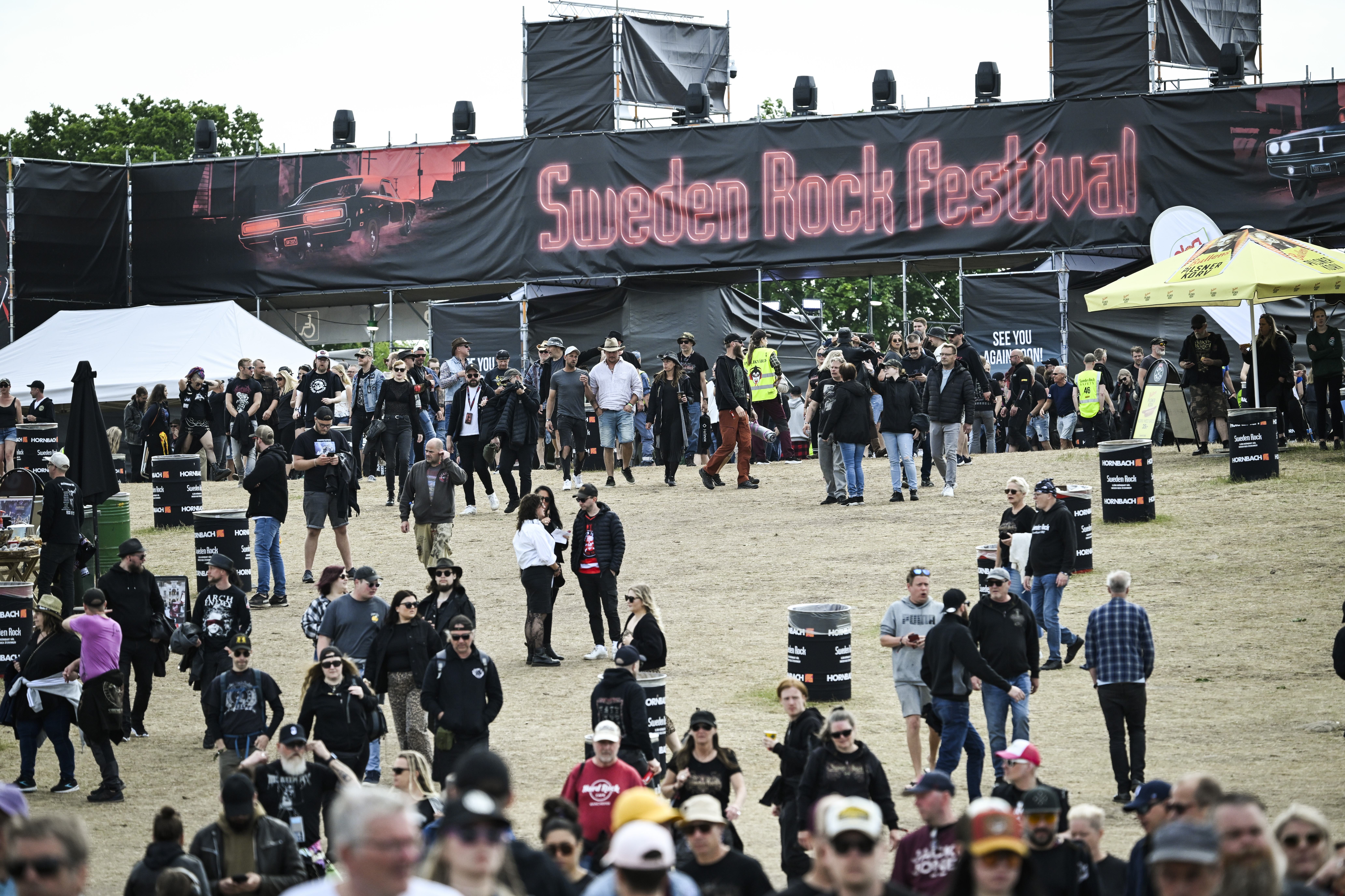 En folkmassa går och samlas utomhus under en skylt med texten "Sweden Rock Festival" på gräs med tält och stånd.