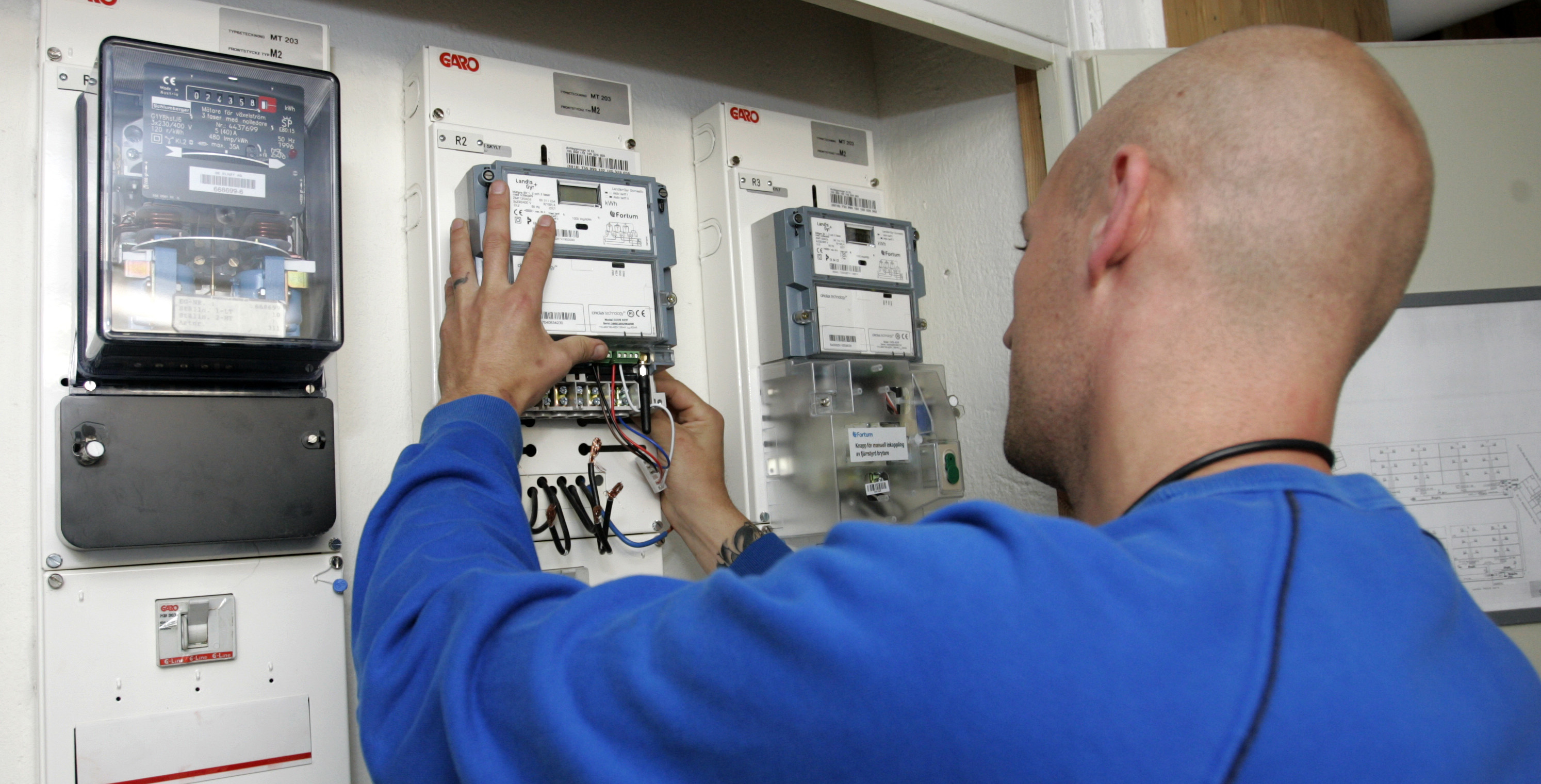 En person med blå skjorta installerar eller inspekterar elmätare på en kontrollpanel.