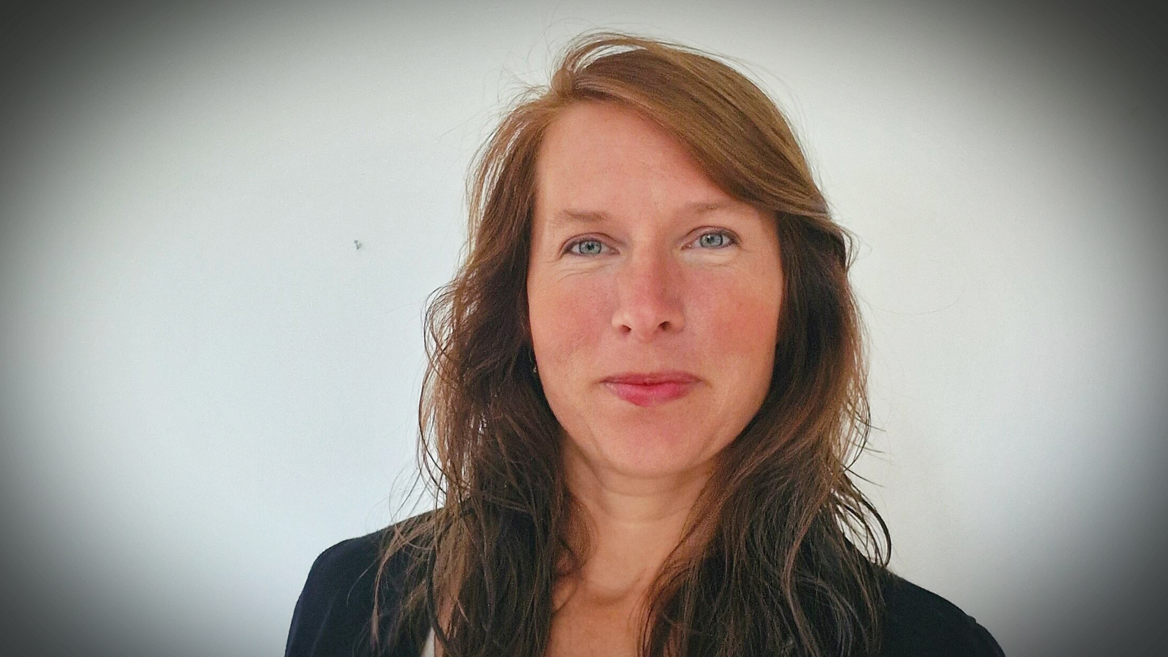 En kvinna med långt brunt hår och blå ögon står framför en vit bakgrund och tittar in i kameran.