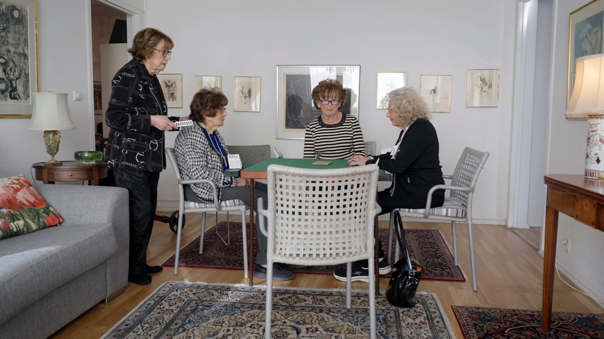 Fyra äldre kvinnor samlas runt ett grönt bord i ett vardagsrum, tre sitter och en står, med konst och mattor runtom.