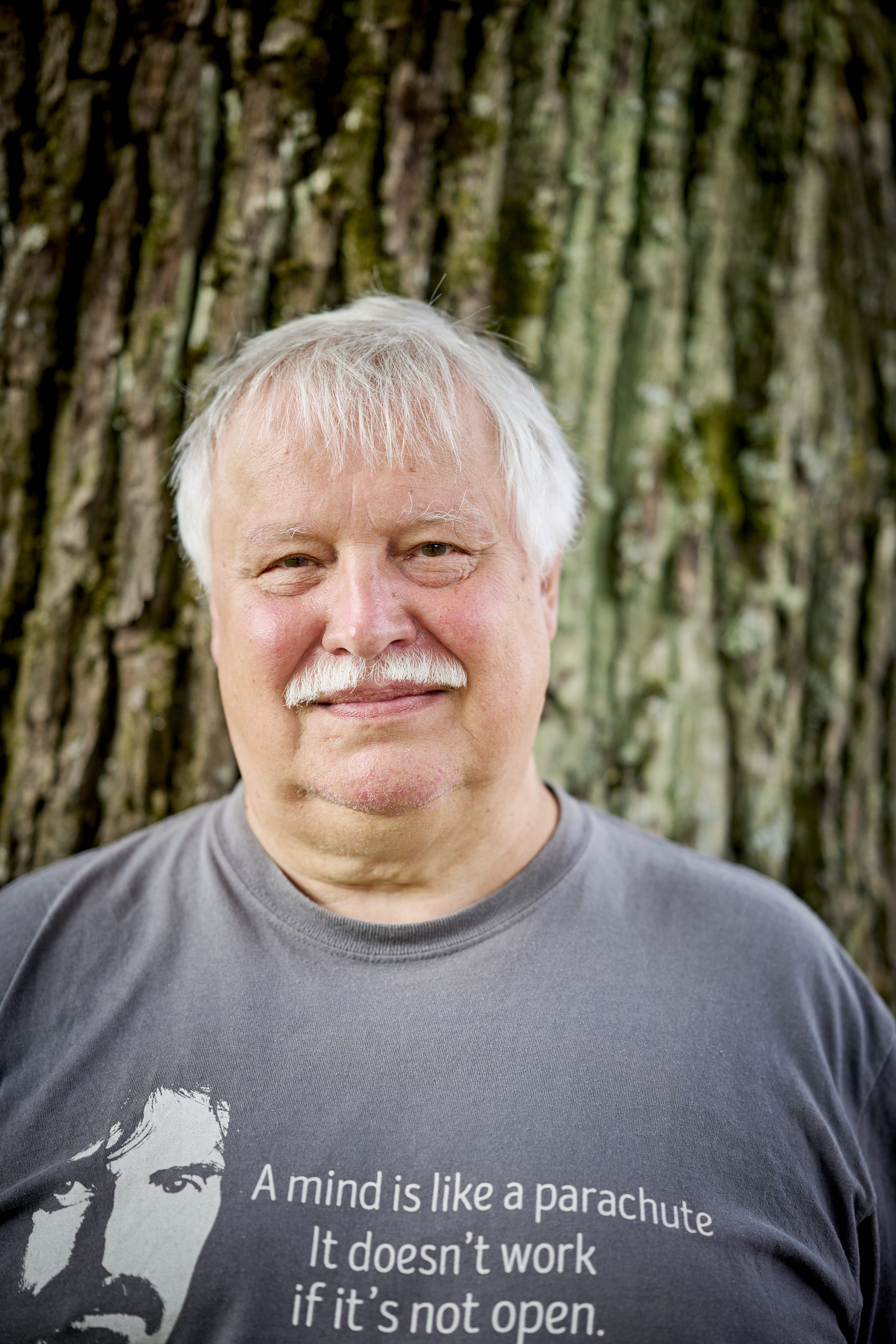 Anders Forslund med vitt hår och mustasch står framför ett träd och har på sig en grå tröja.