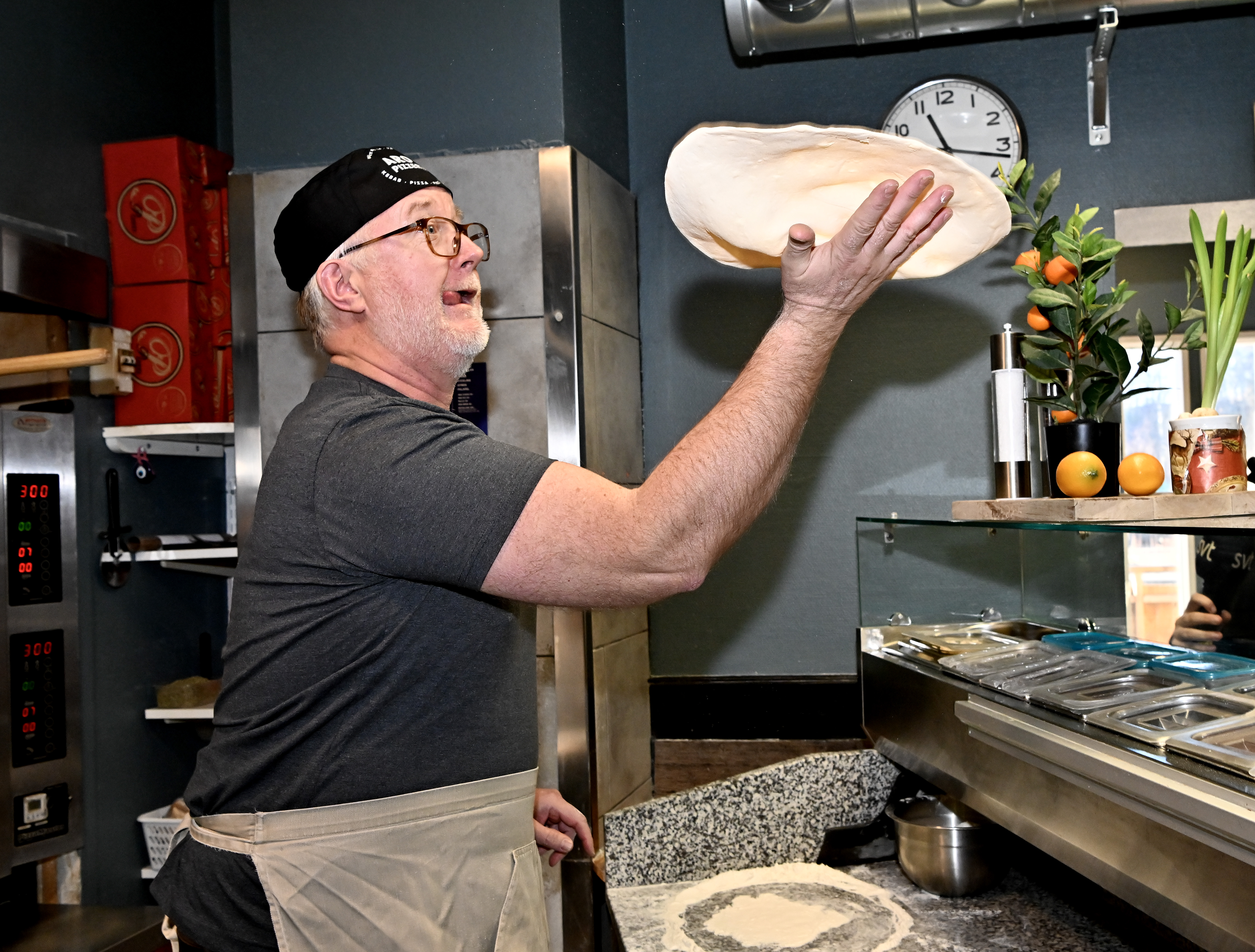 Johan Pehrson med svart keps och förkläde slungar pizzadeg i luften i ett kök, med en disk och ingredienser intill.