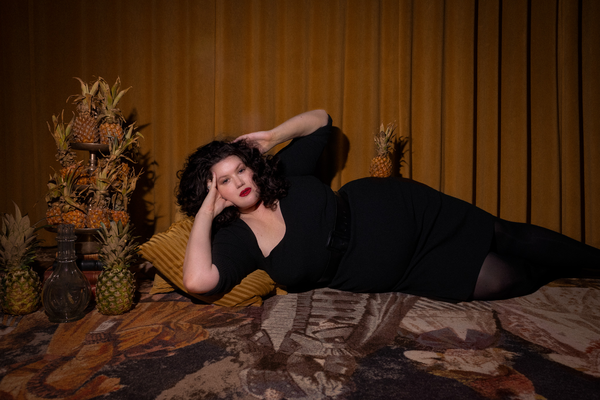 En person i svart klänning ligger på en kudde framför ett gyllene draperi, med ananas och karaffer bredvid sig.