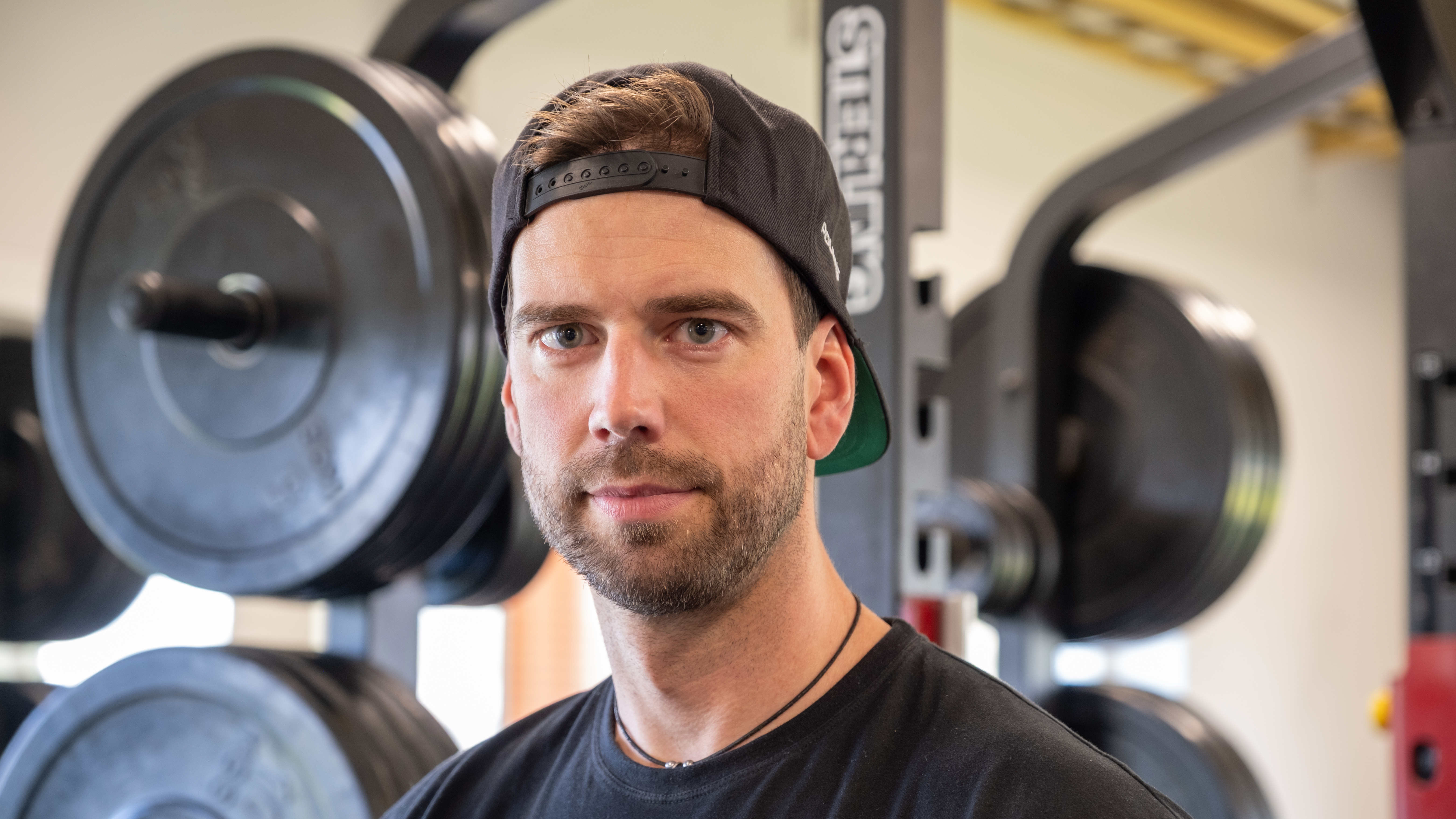 En man med svart t-shirt och bakåtvänd keps står framför styrketräningsutrustning i ett gym.
