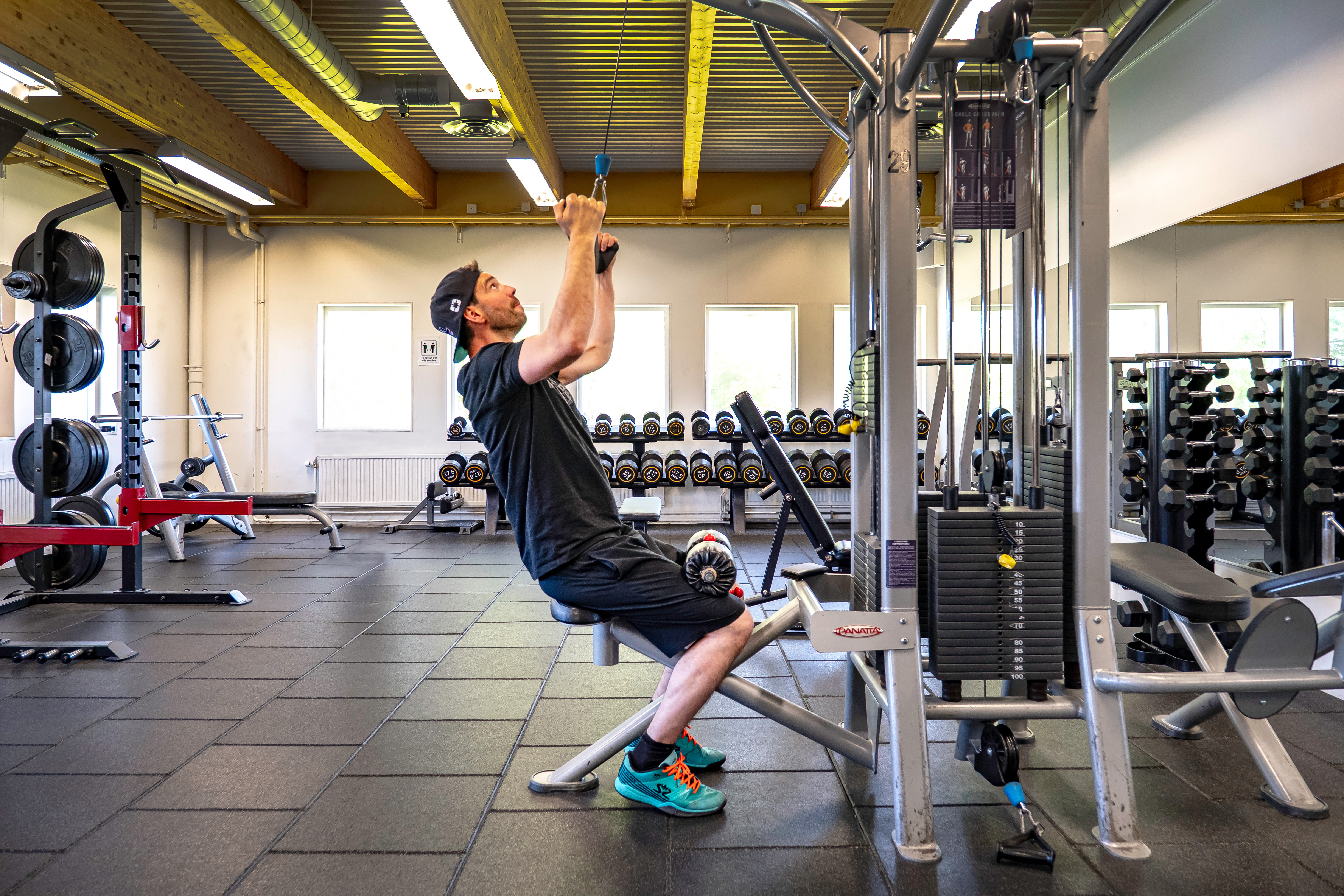 En man i träningskläder använder en latsdragsmaskin i ett gym med vikter och annan träningsutrustning.