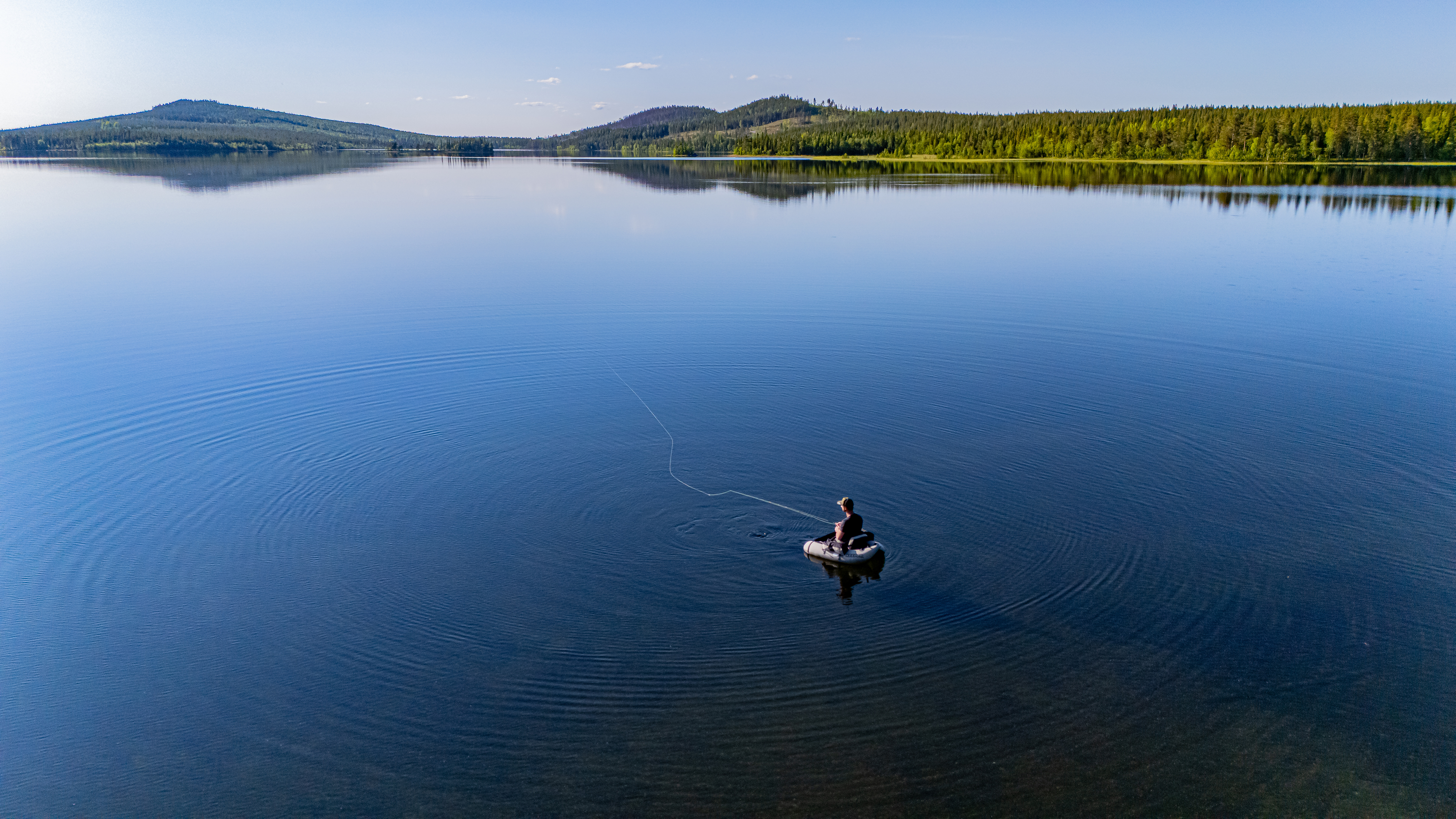 En person sitter i en liten uppblåsbar båt och fiskar på en sjö omgiven av skogsklädda kullar under en blå himmel.