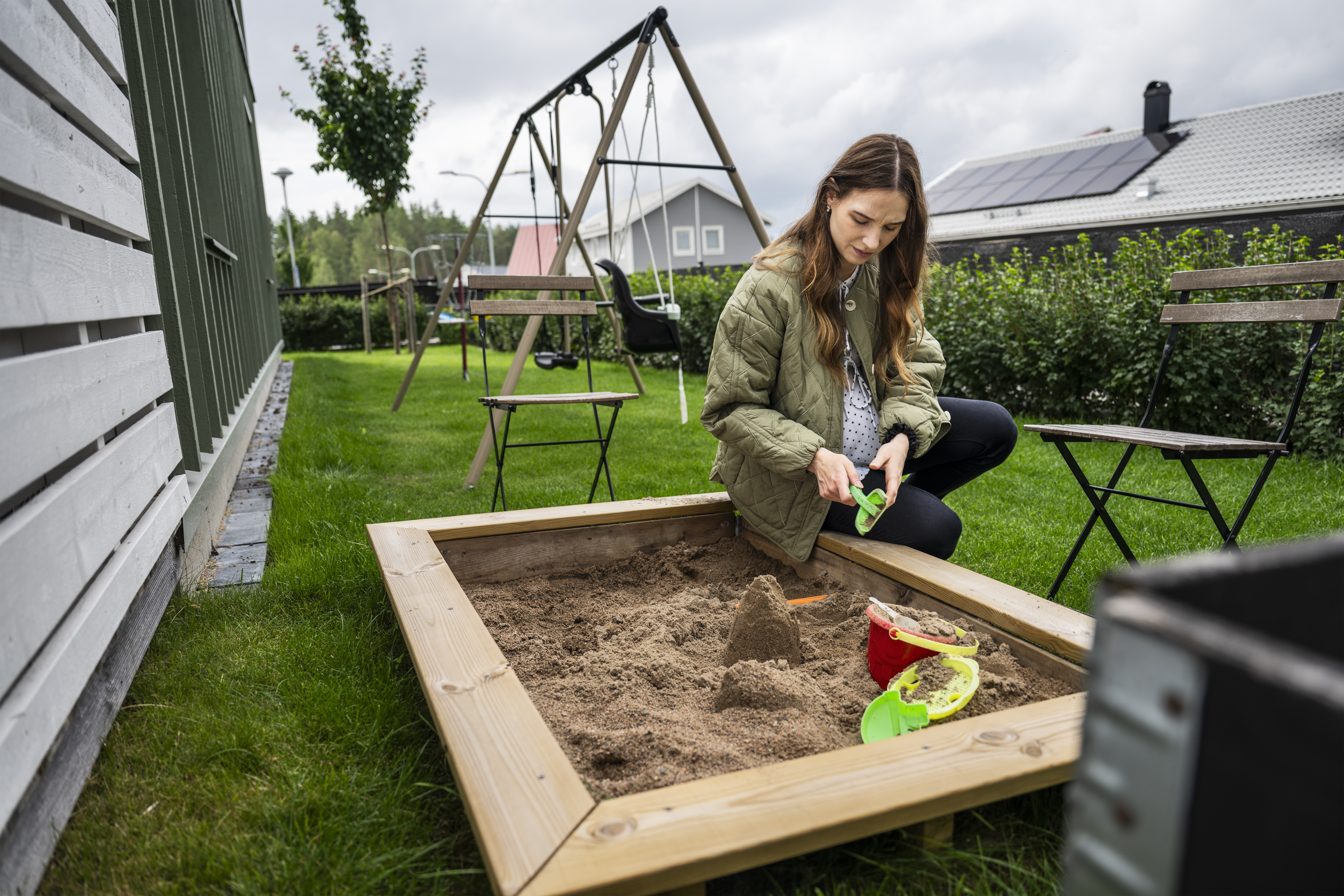 Kristina Lindholm sitter på knä vid en sandlåda i en trädgård och leker med sand med en grön spade. I bakgrunden finns stolar, en gunga och hus.