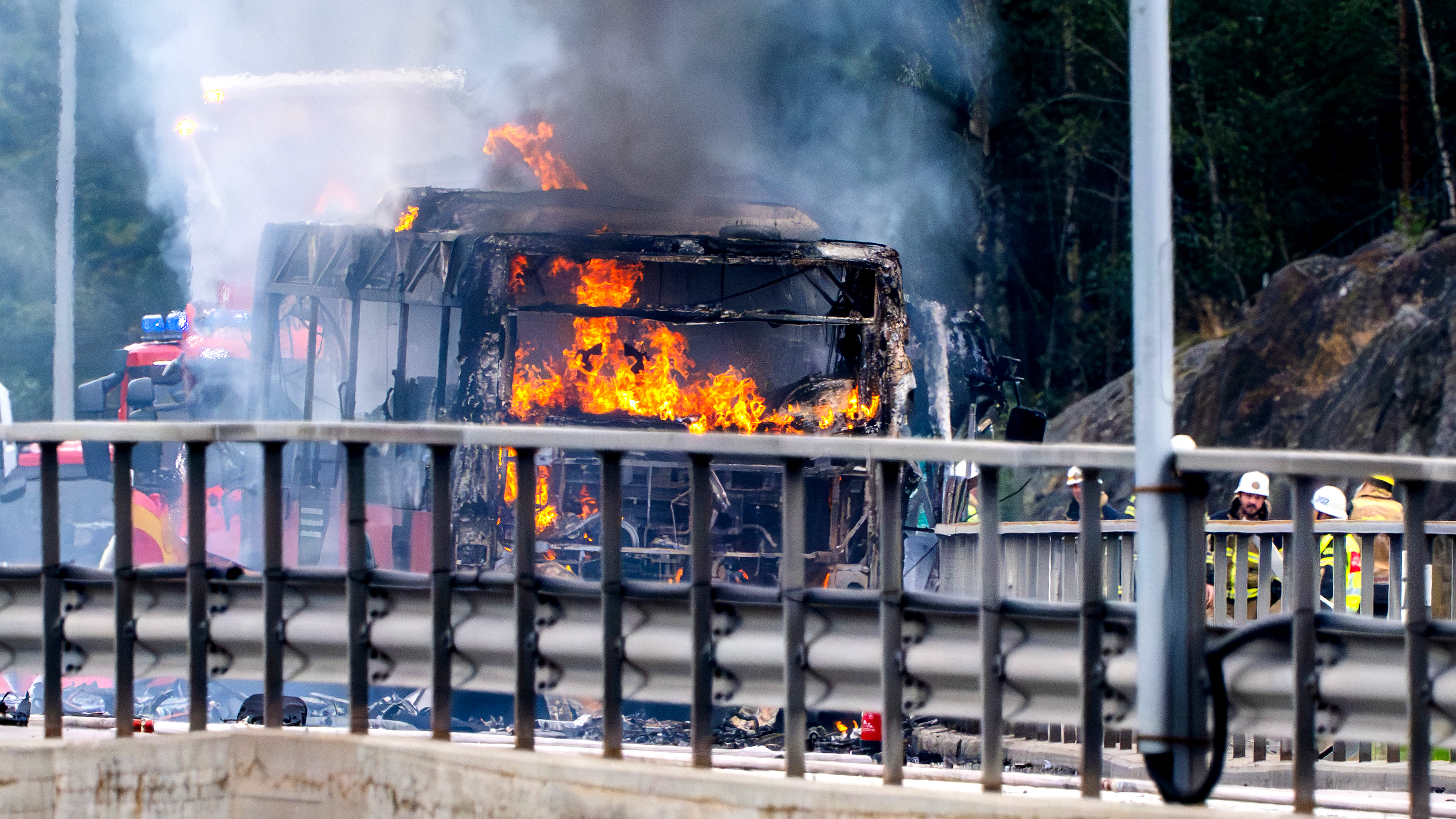 En buss brinner på en väg, rök syns. Brandmän och räddningspersonal står bakom ett avspärrningsband i närheten.