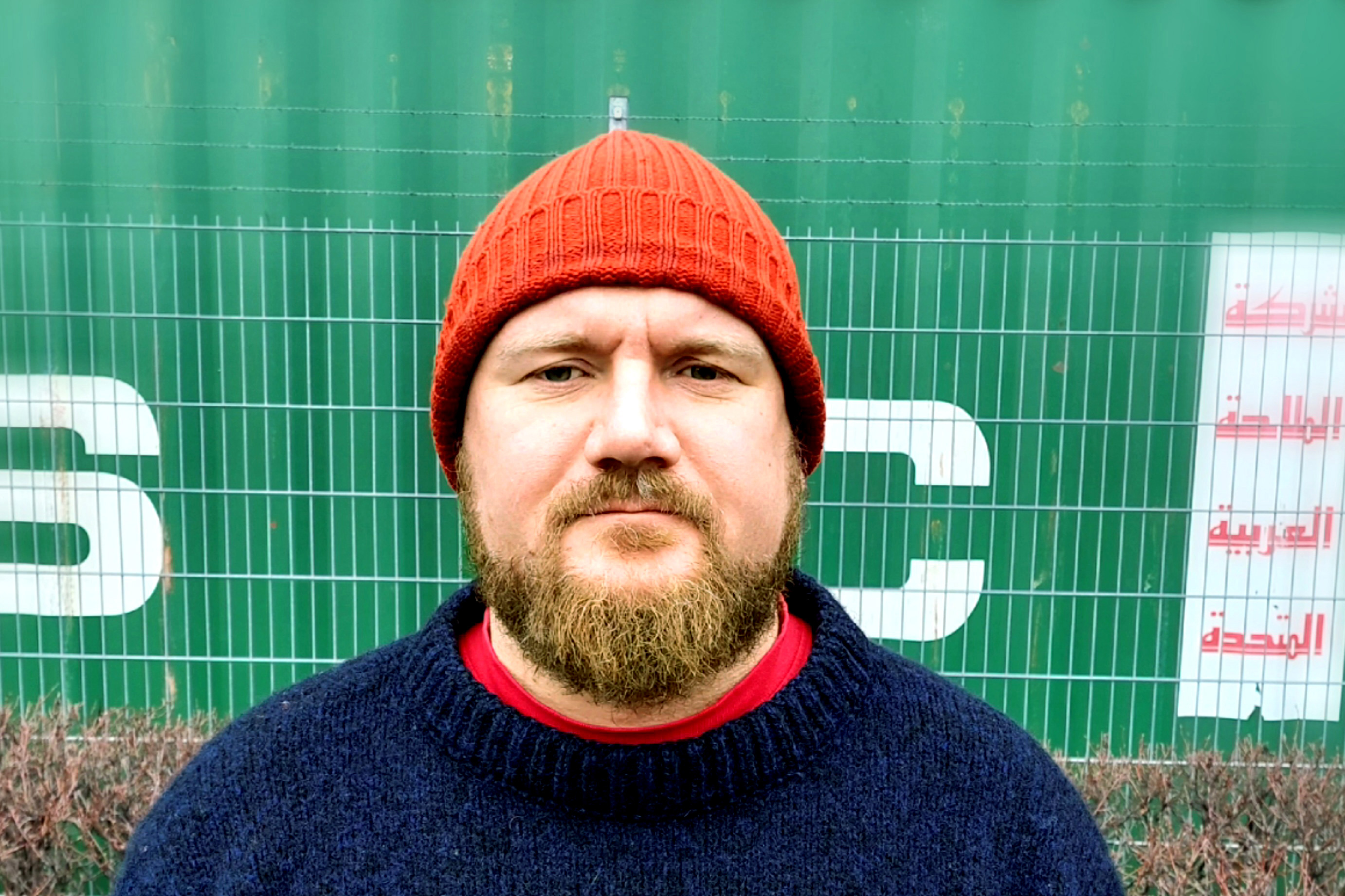 Skäggig man med röd stickad mössa och mörk tröja står framför ett grönt stängsel och en fraktcontainer med bokstäver och arabisk text.