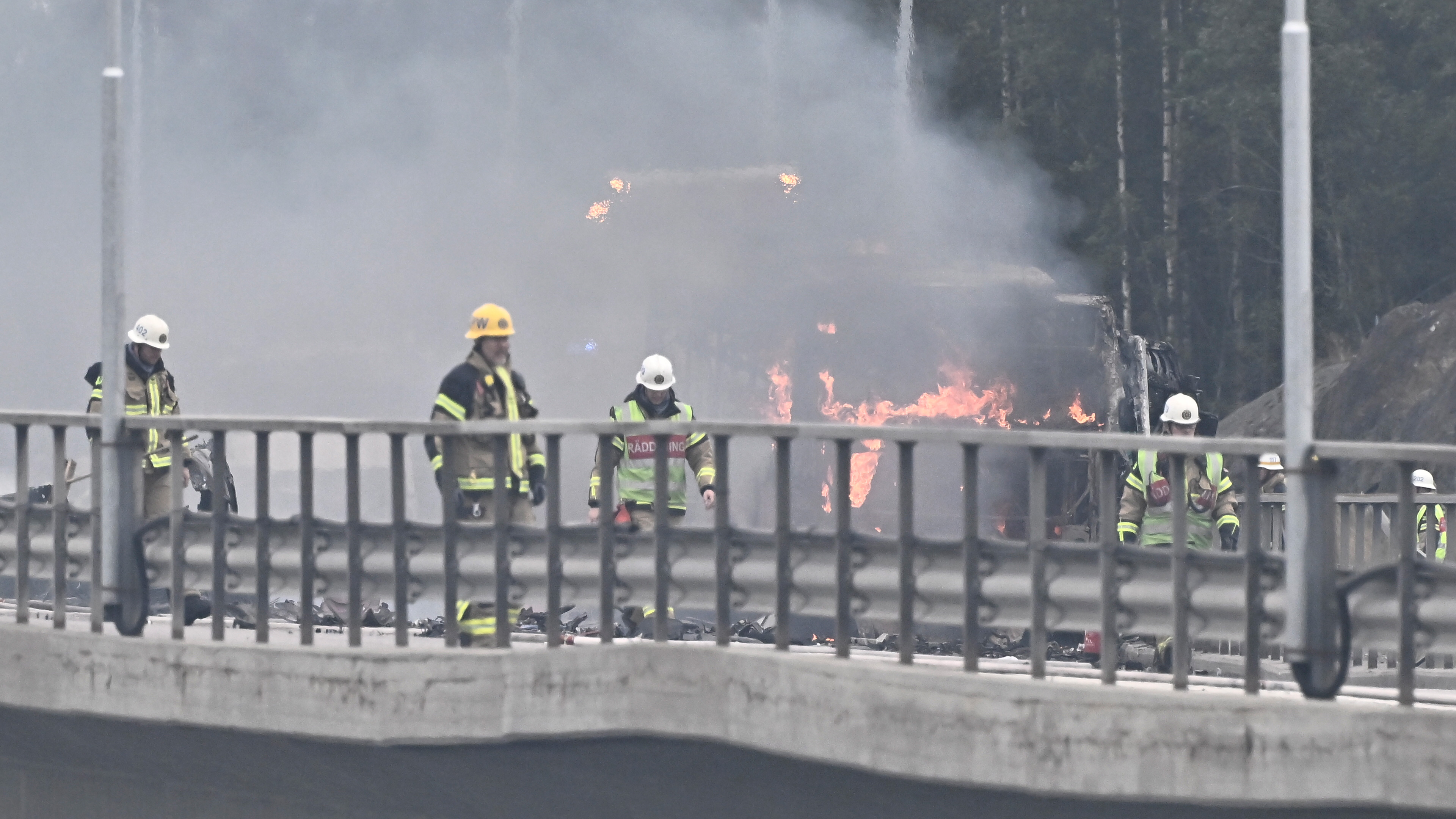 Brandmän arbetar vid en bilbrand på en motorväg. Rök och eld syns i bakgrunden.