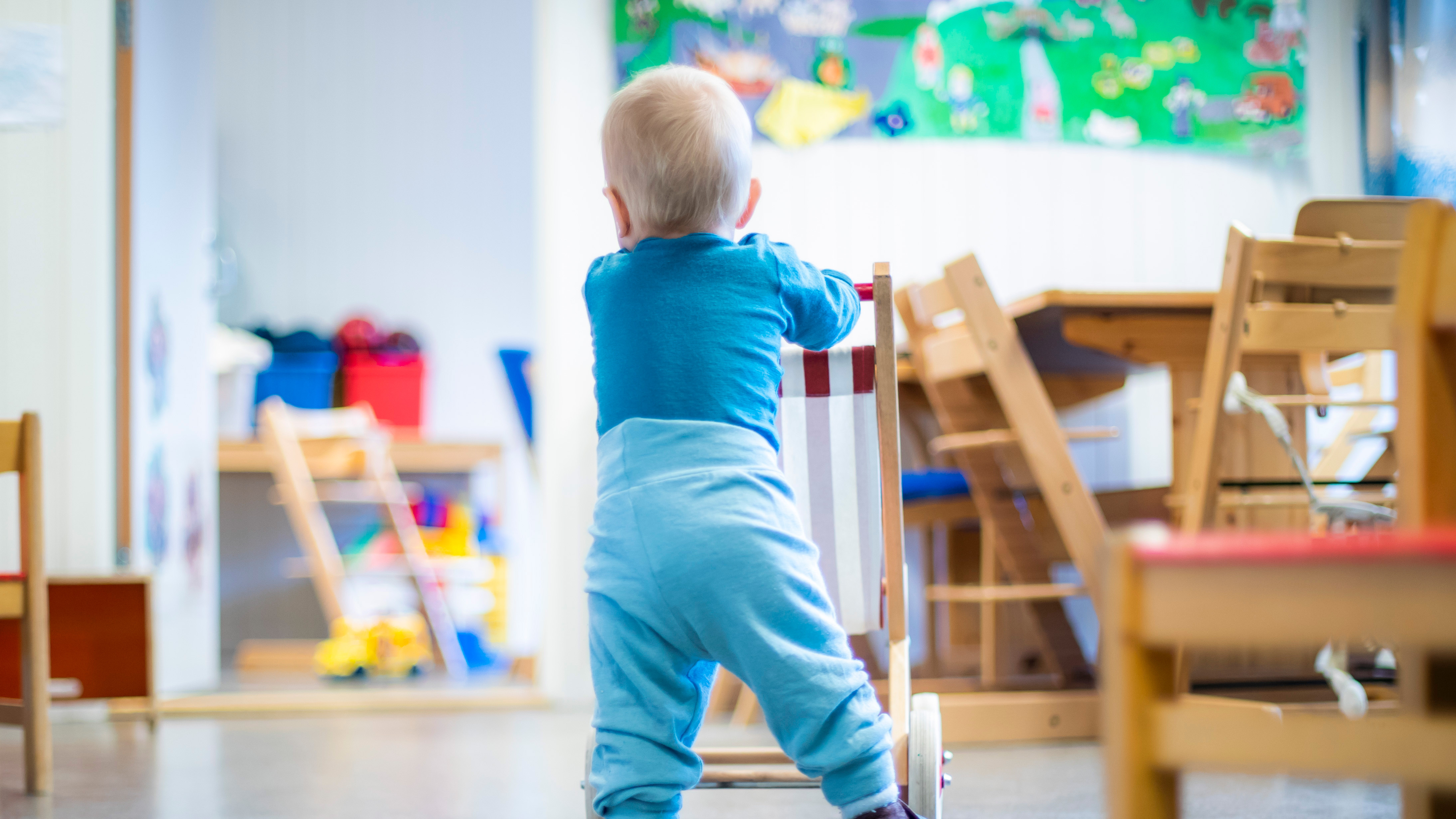 En baby i blå kläder står och skjuter en träläragång i ett rum med barnmöbler och färgglada väggdekorationer.