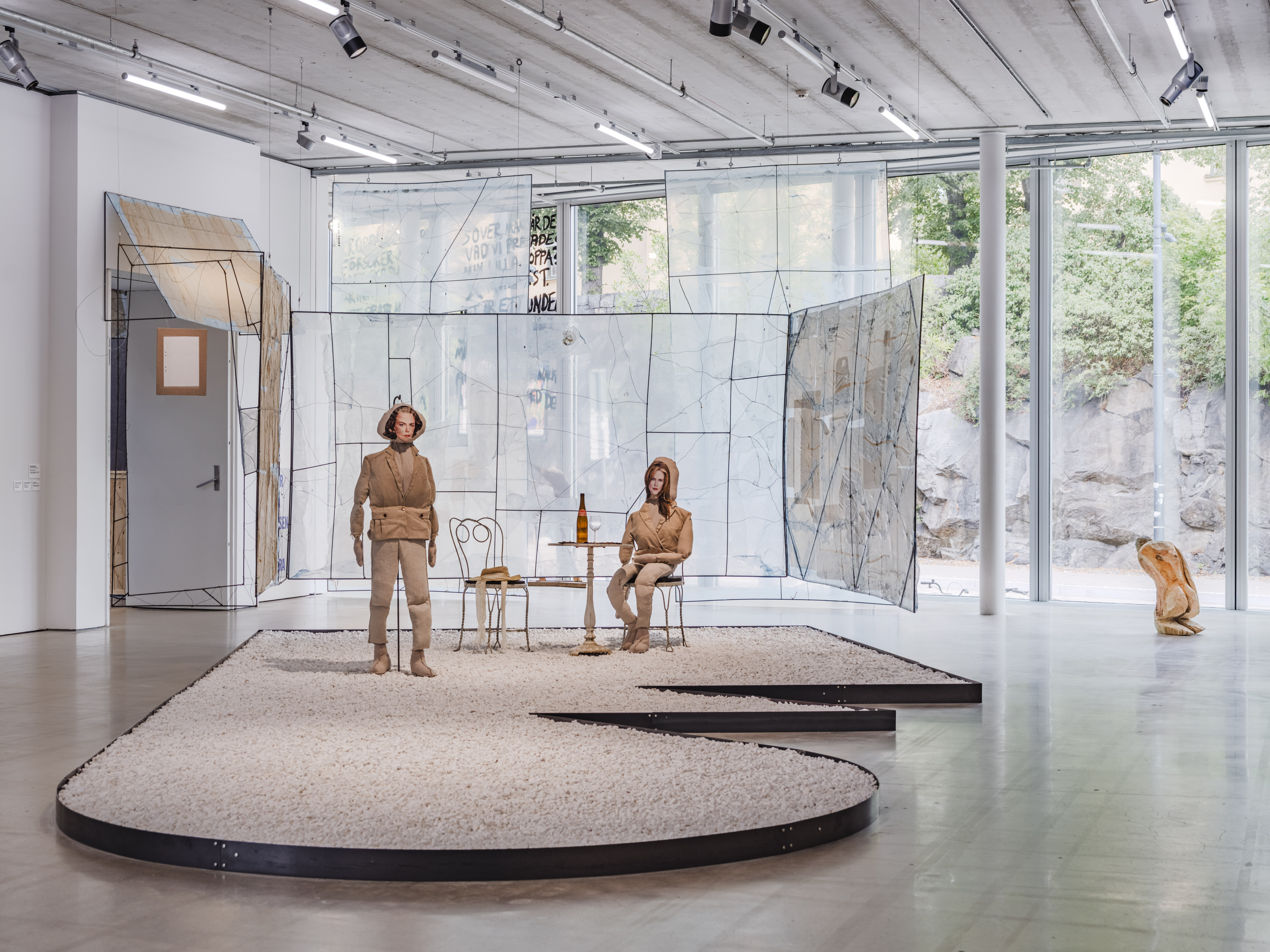 Två människoliknande figurer i beige står och sitter på en grusplattform i ett galleri med glas- och metallväggar.