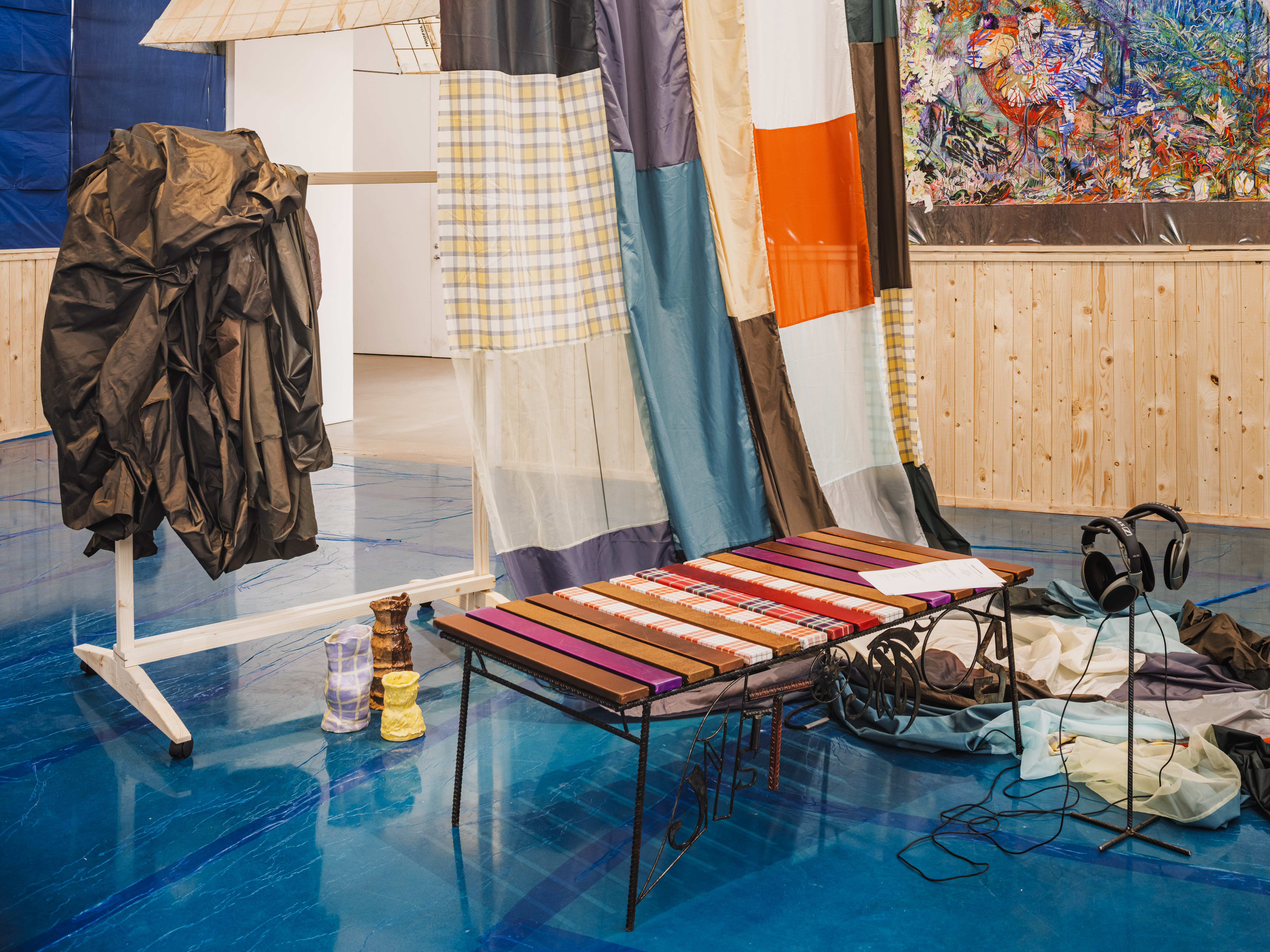 Ett rum med blått golv, lapptäcksgardiner, en randig bänk, två par hörlurar, kläder på ett ställ och konst på trävägg.